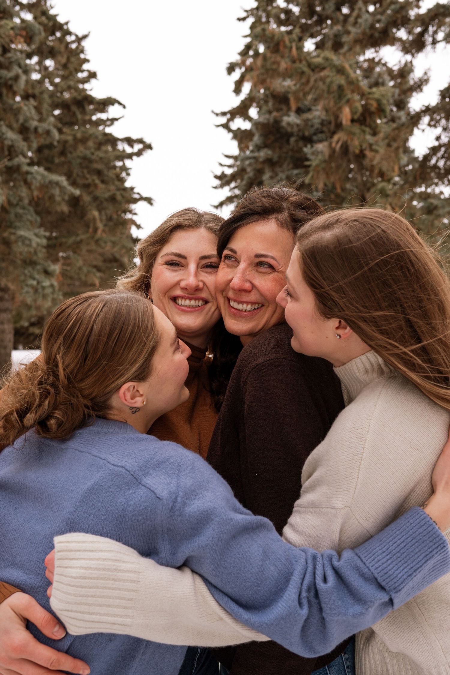 Four women hugging and smiling outdoors in winter, surrounded by snow-dusted evergreen trees.