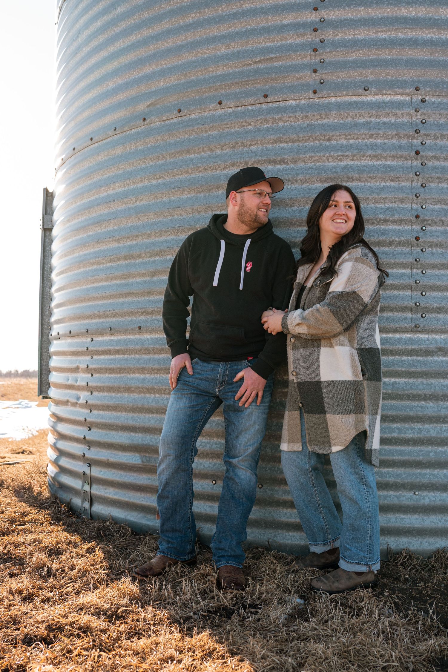 Couple smiling together leaning against a rustic grain silo on a farm during golden hour.