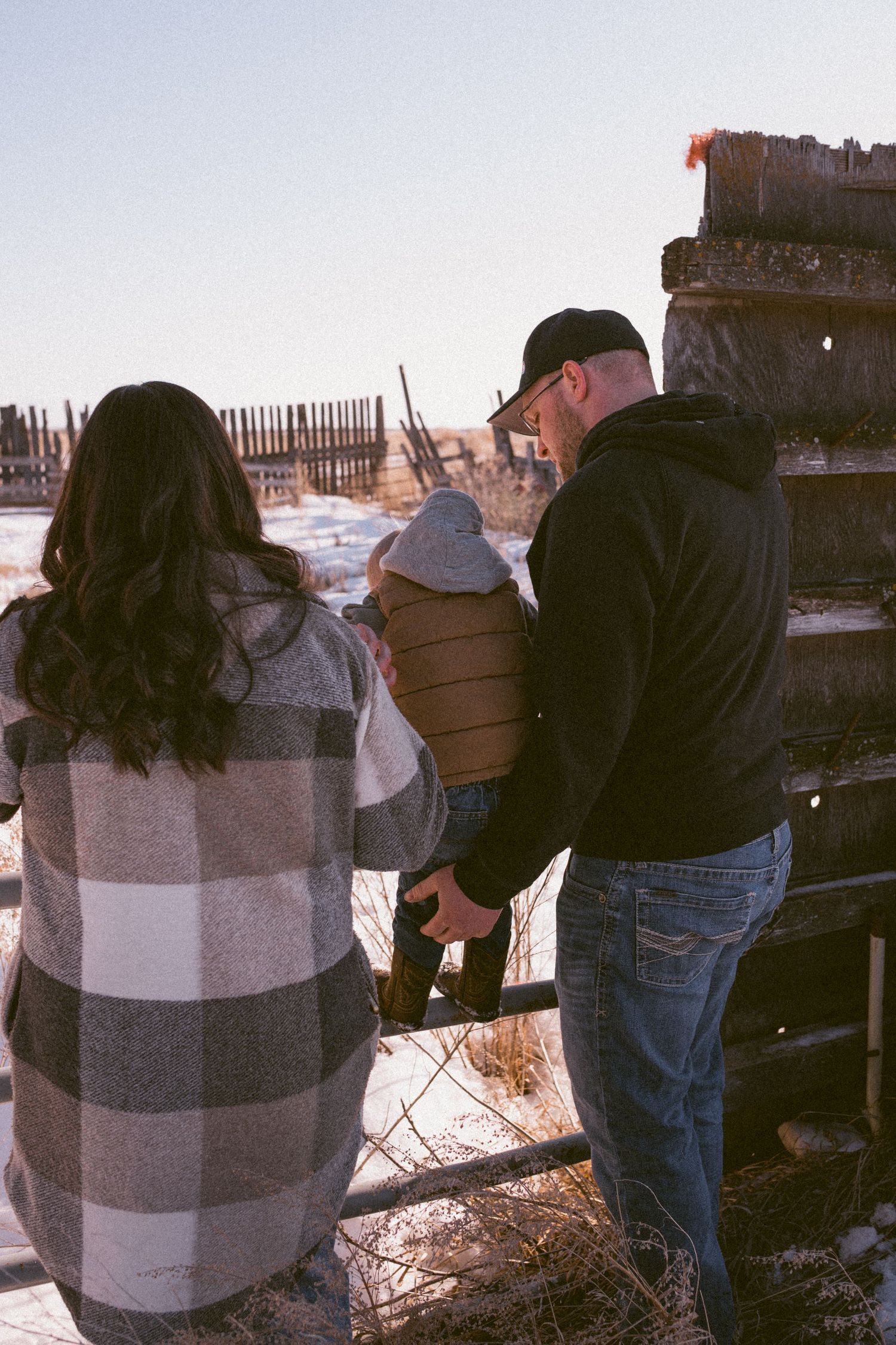 Family of three holding hands at a snowy winter fence, viewed from behind, wearing plaid and black hoodie.