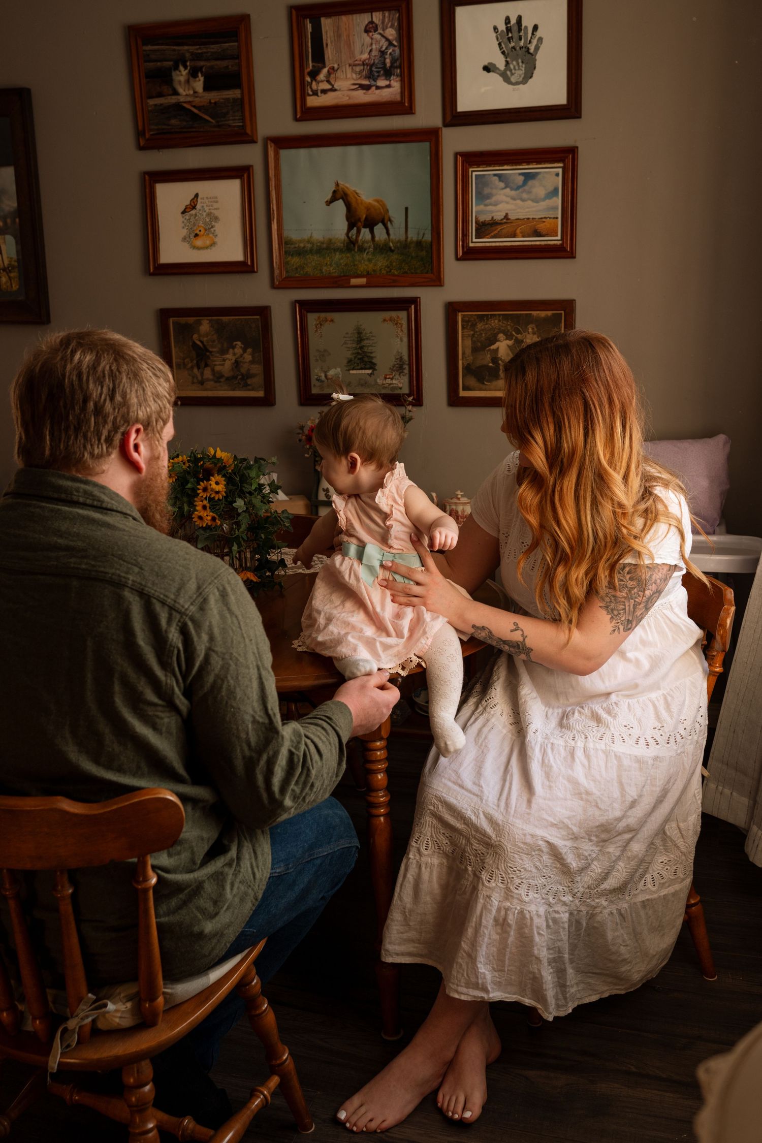 A family sits at a wooden table admiring their baby girl, surrounded by framed artwork on a warm, dimly lit wall.