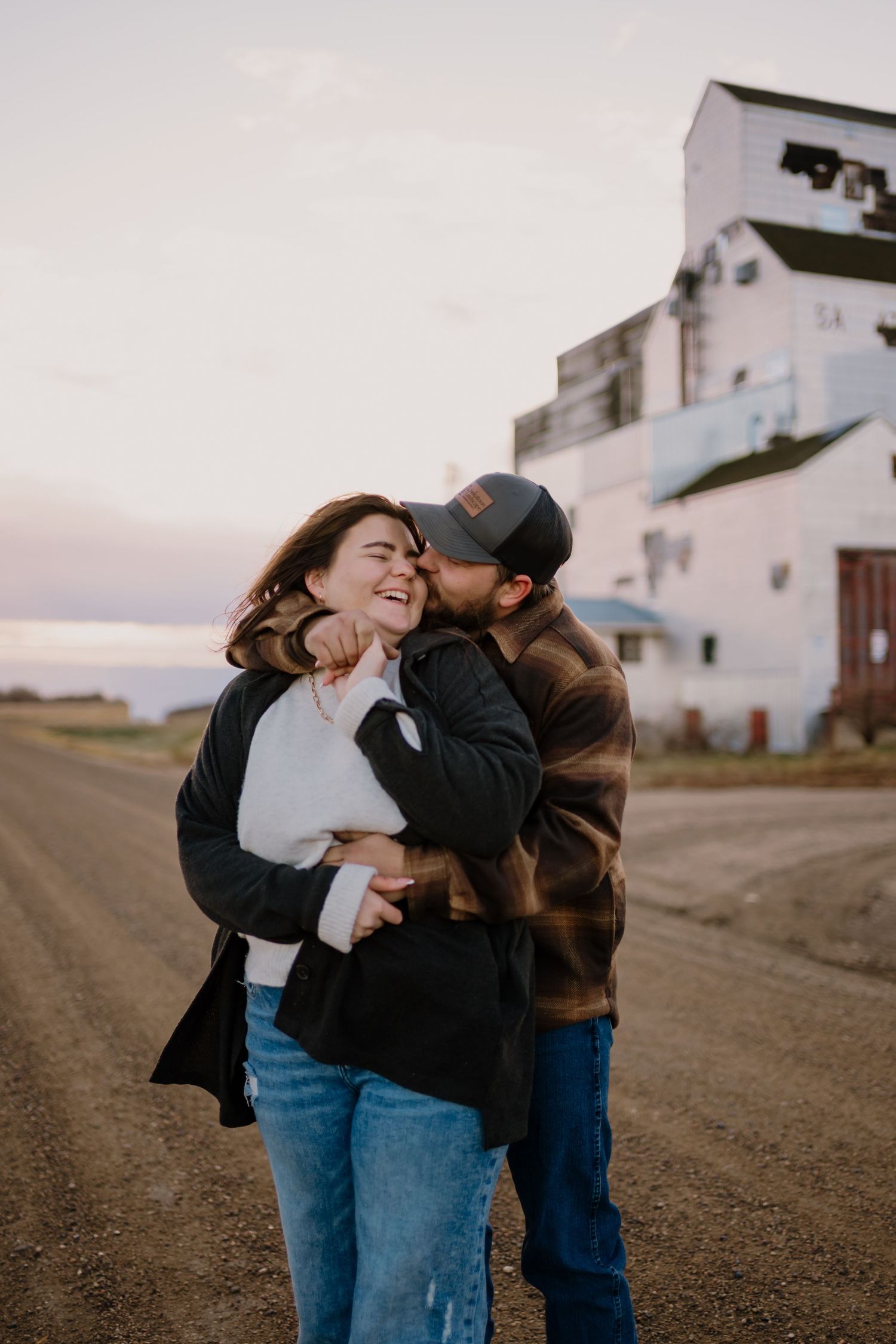 A couple shares an intimate moment on a dirt road at dusk with buildings in the background.