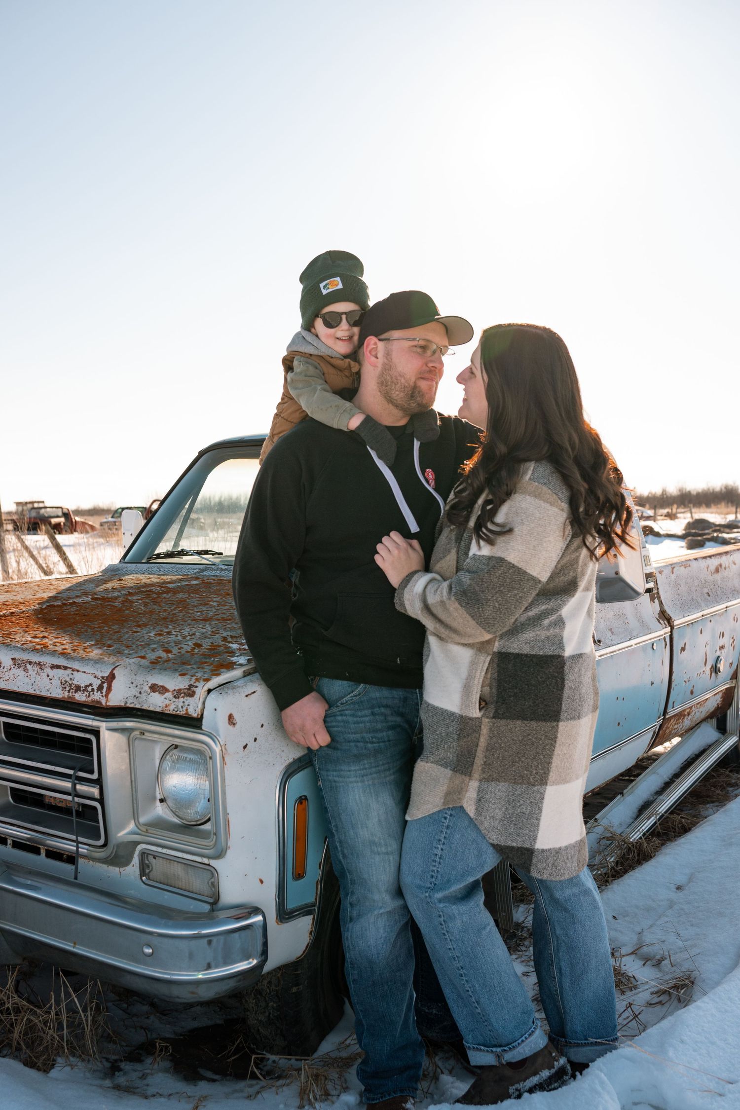A couple and child pose by a rustic old truck in a snowy winter field, dressed in casual cold-weather attire.