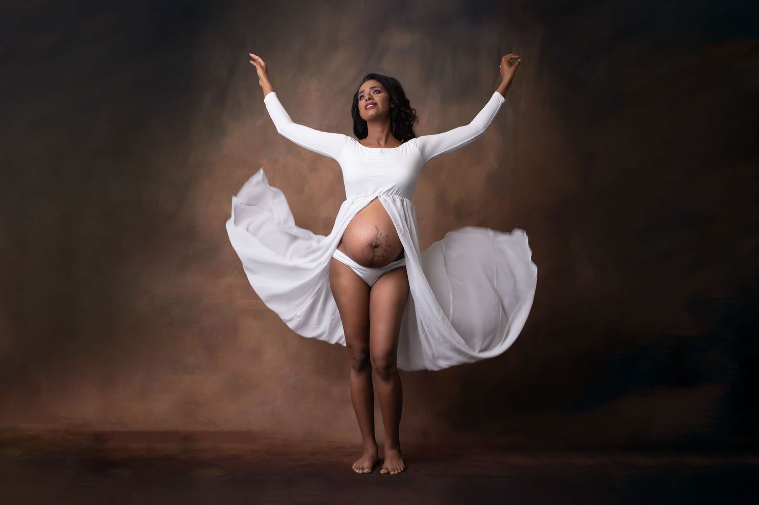 Pregnant woman in flowing white dress posing gracefully with arms raised in a professional maternity photo studio.