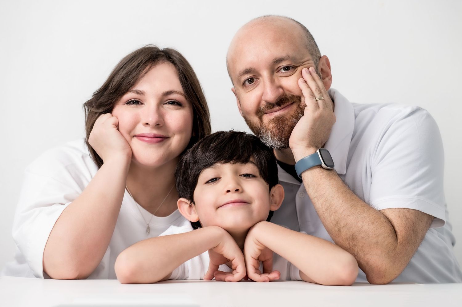 A smiling family of three poses together, with parents resting their chins on their hands beside their young son against a white background.