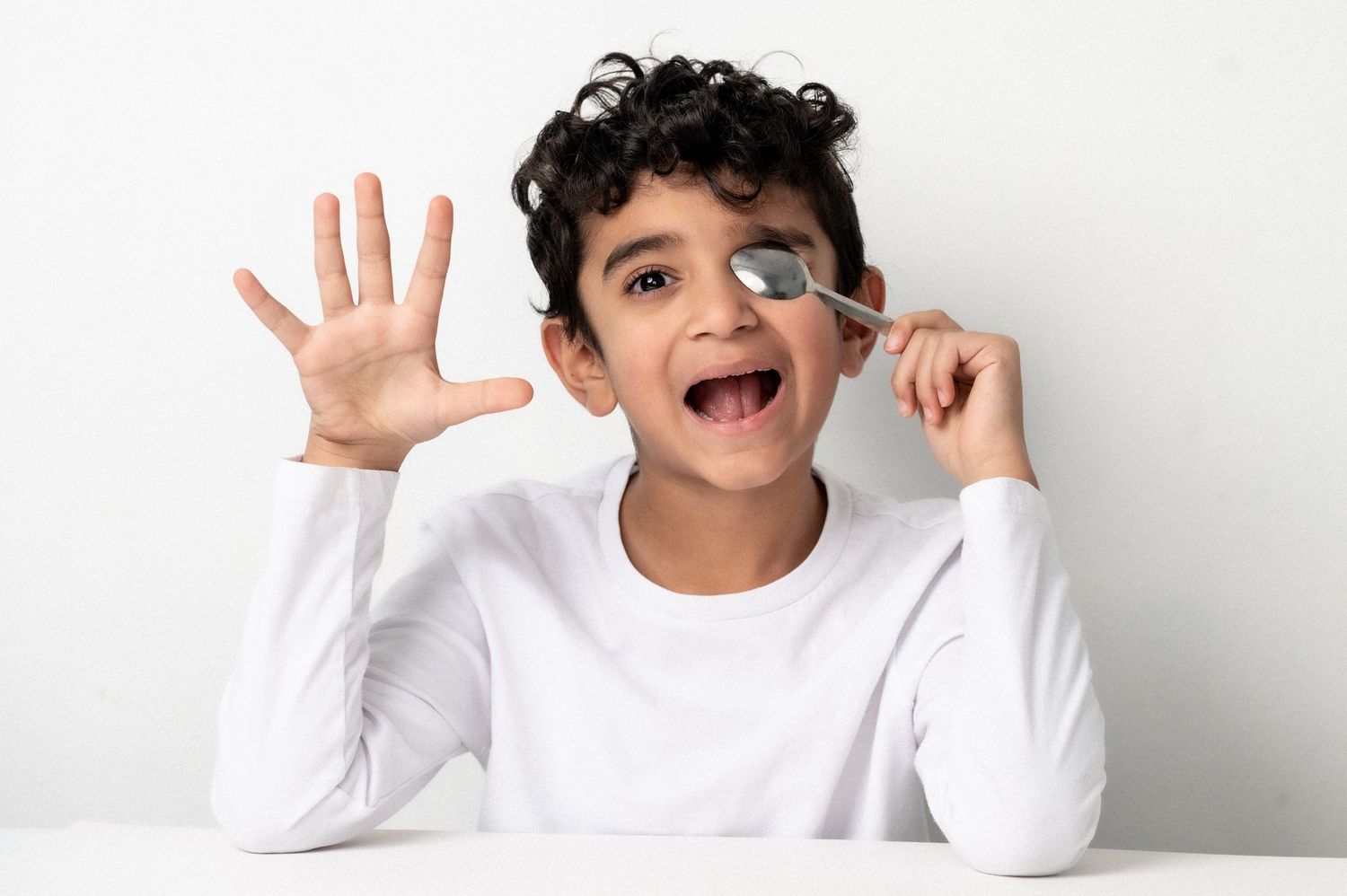 Happy boy holding a spoon near his eye with his hand raised, wearing a white long-sleeve shirt against a white background.