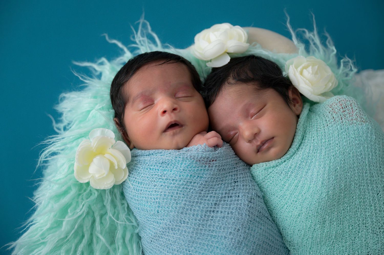 Two sleeping newborn twins wrapped in teal knit blankets with white flower headbands on a turquoise background.