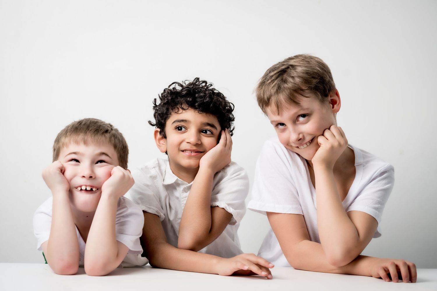 Three young boys in white t-shirts posing with chins on hands, smiling at camera against white background.