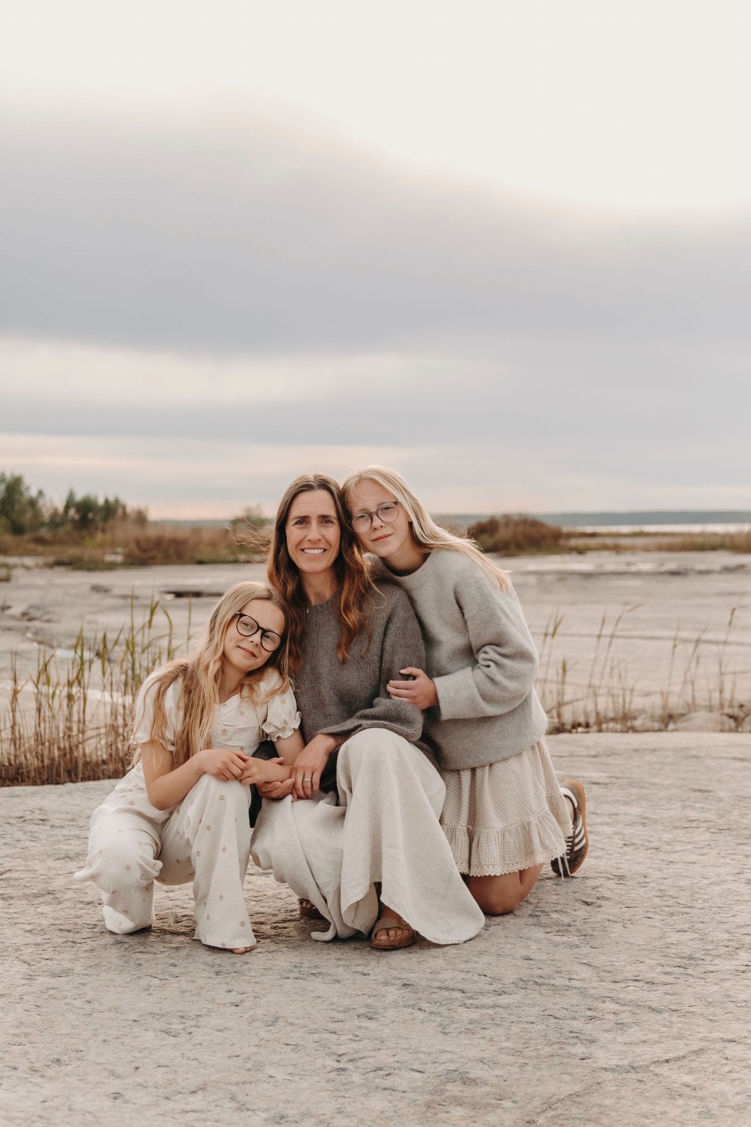 Three people in neutral tones sit together on a beach with grasses and coastal views on an overcast day.