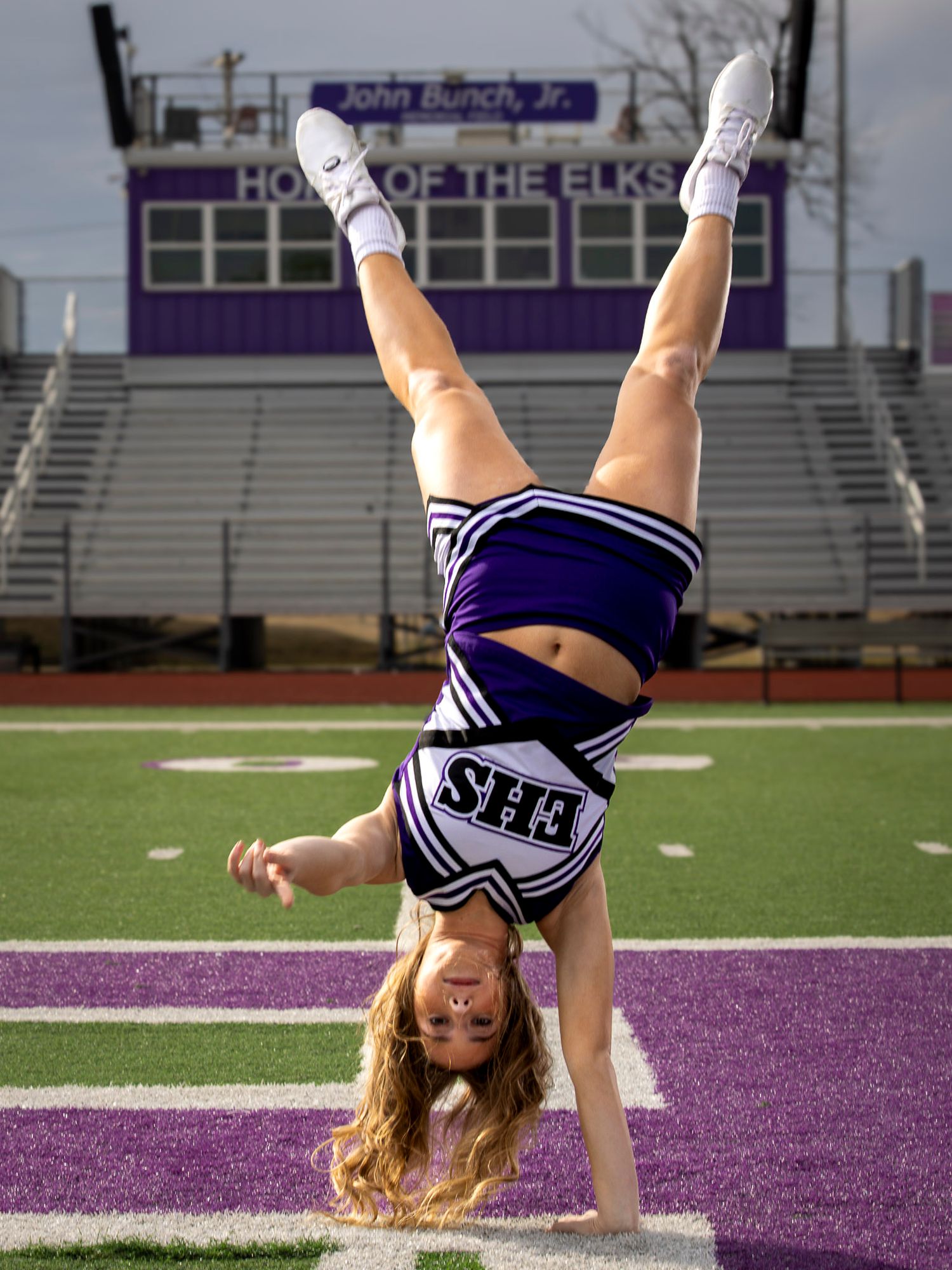 Cheerleader performing a one-handed cartwheel on a purple football field in front of stadium bleachers.