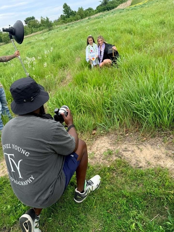 Photographer in TY Photography shirt captures two people posing in a lush green field during an outdoor photo shoot.
