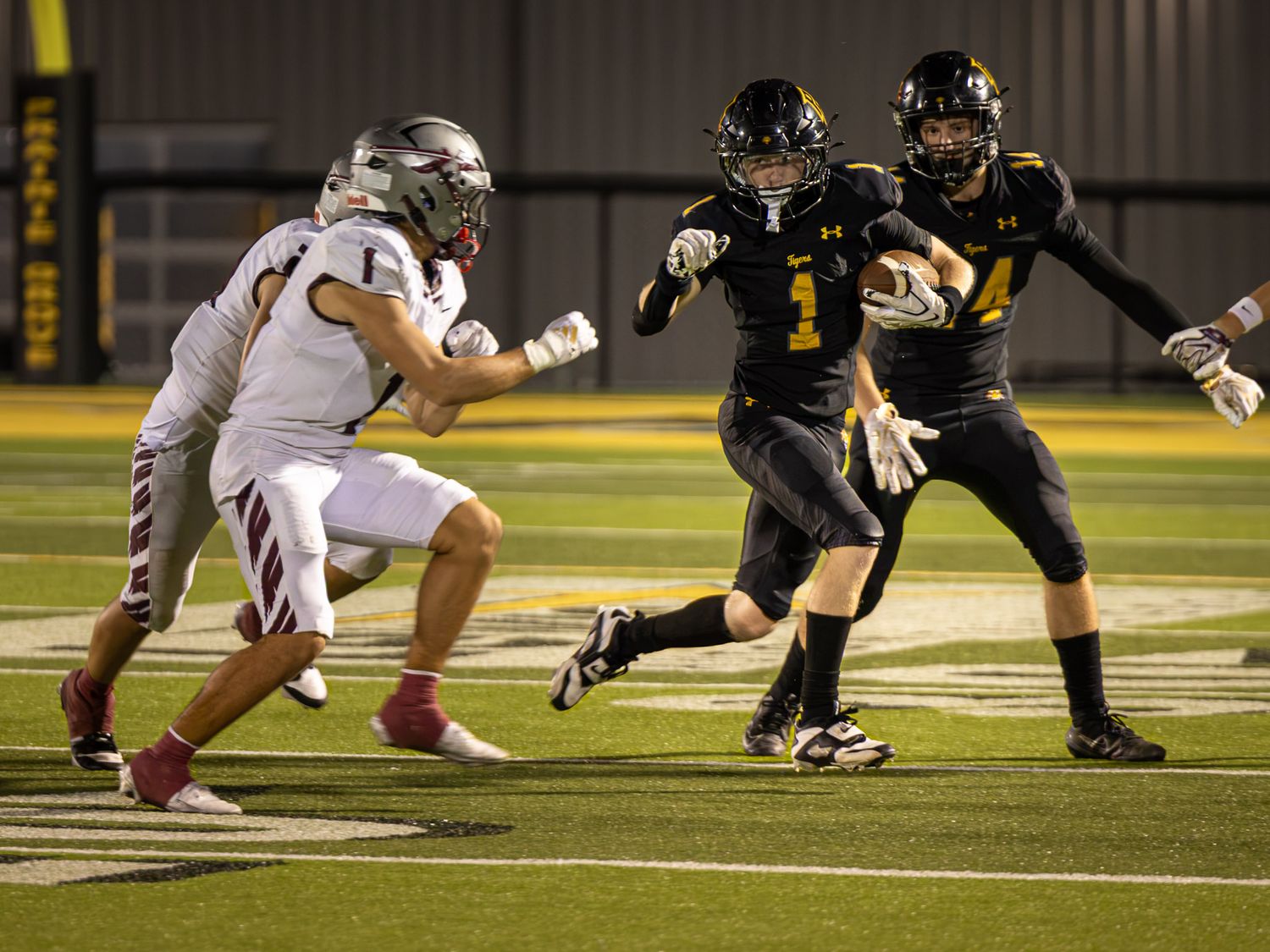 Football player in black and gold uniform number 1 carries the ball past a defender in white during a night game.