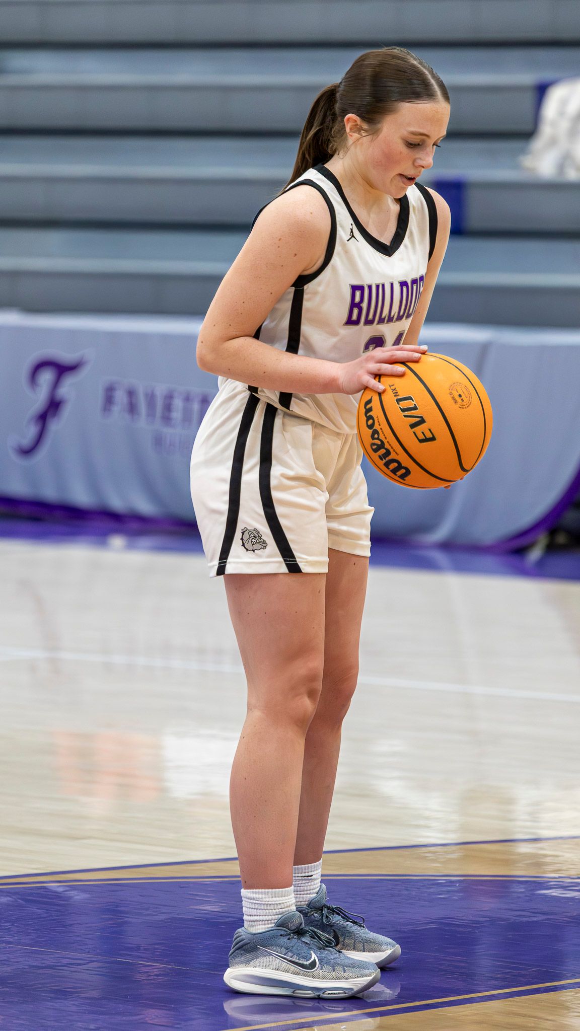 Basketball player in white Bulldogs uniform stands on purple court holding a basketball during game preparation.