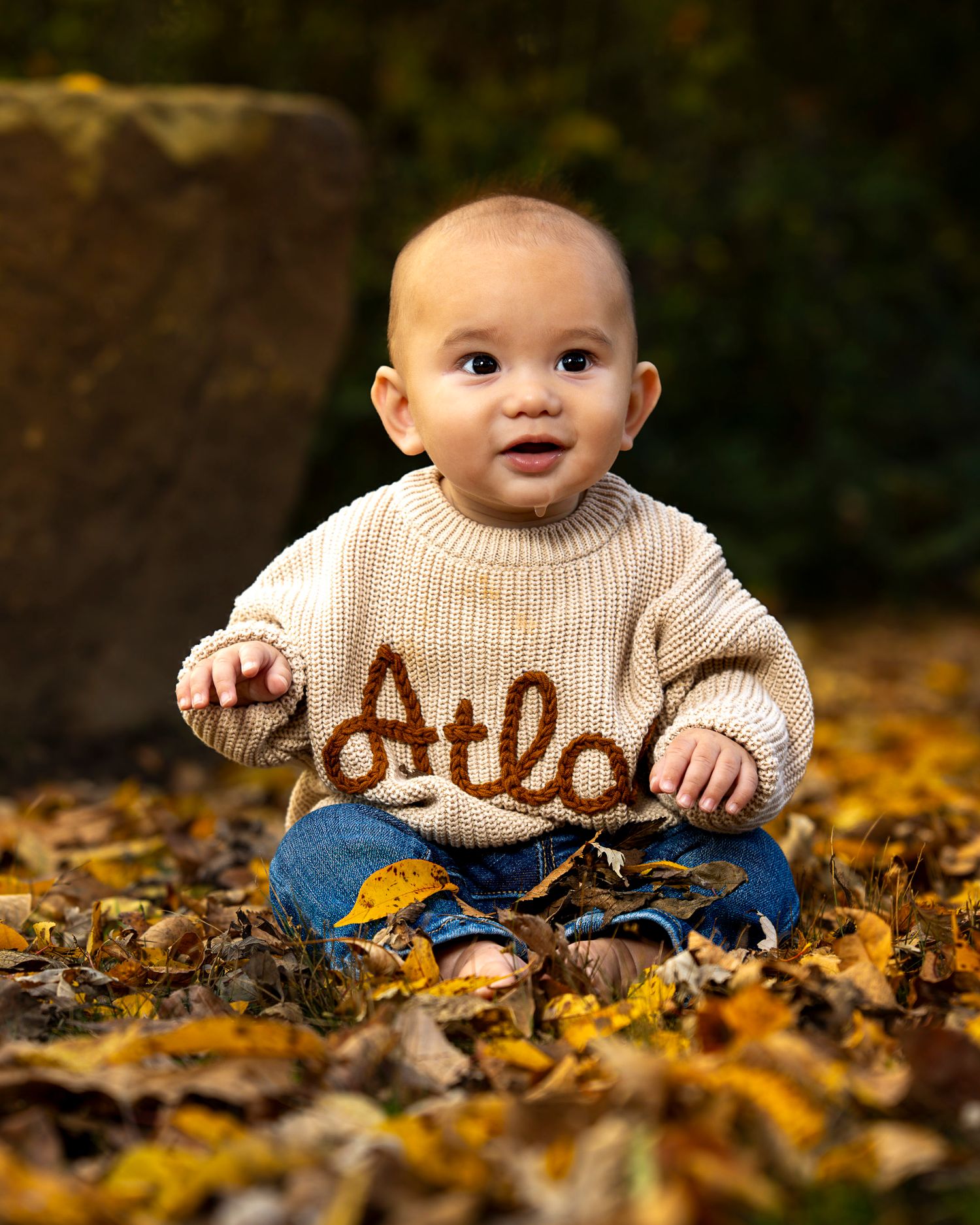 Baby in beige sweater sits among fallen autumn leaves on the ground.