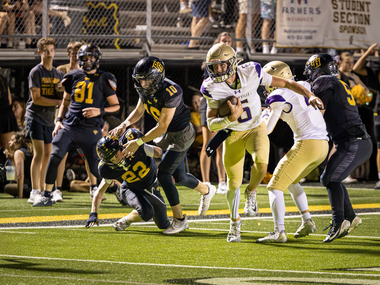 High school football player in a white jersey carries the ball past diving defenders in dark uniforms during a Friday night game.