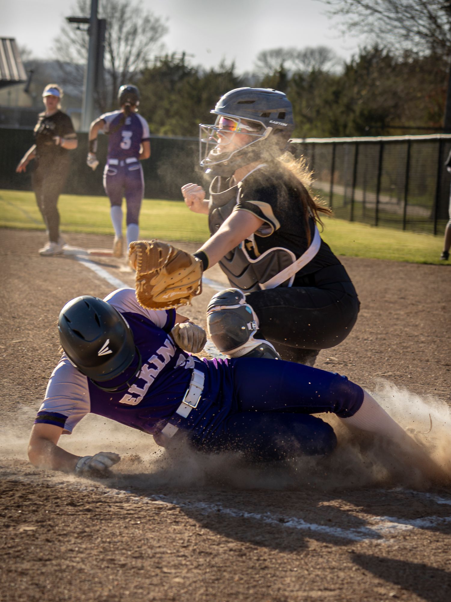 A softball runner in a purple uniform slides into home plate in a cloud of dust as the catcher reaches with her glove for the tag.