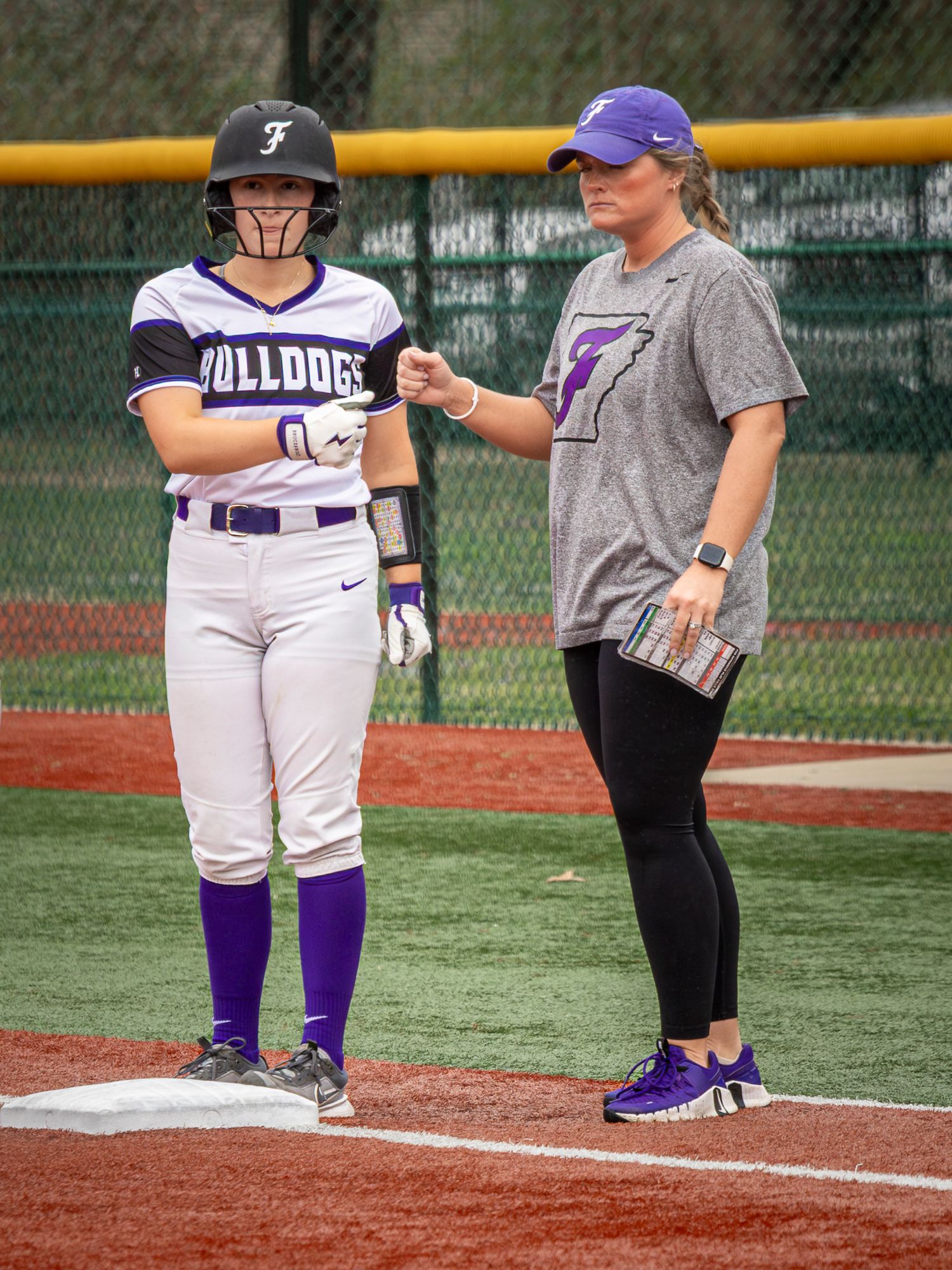 Bulldogs softball player in a batting helmet stands on third base while a coach in a purple cap gives her instructions.