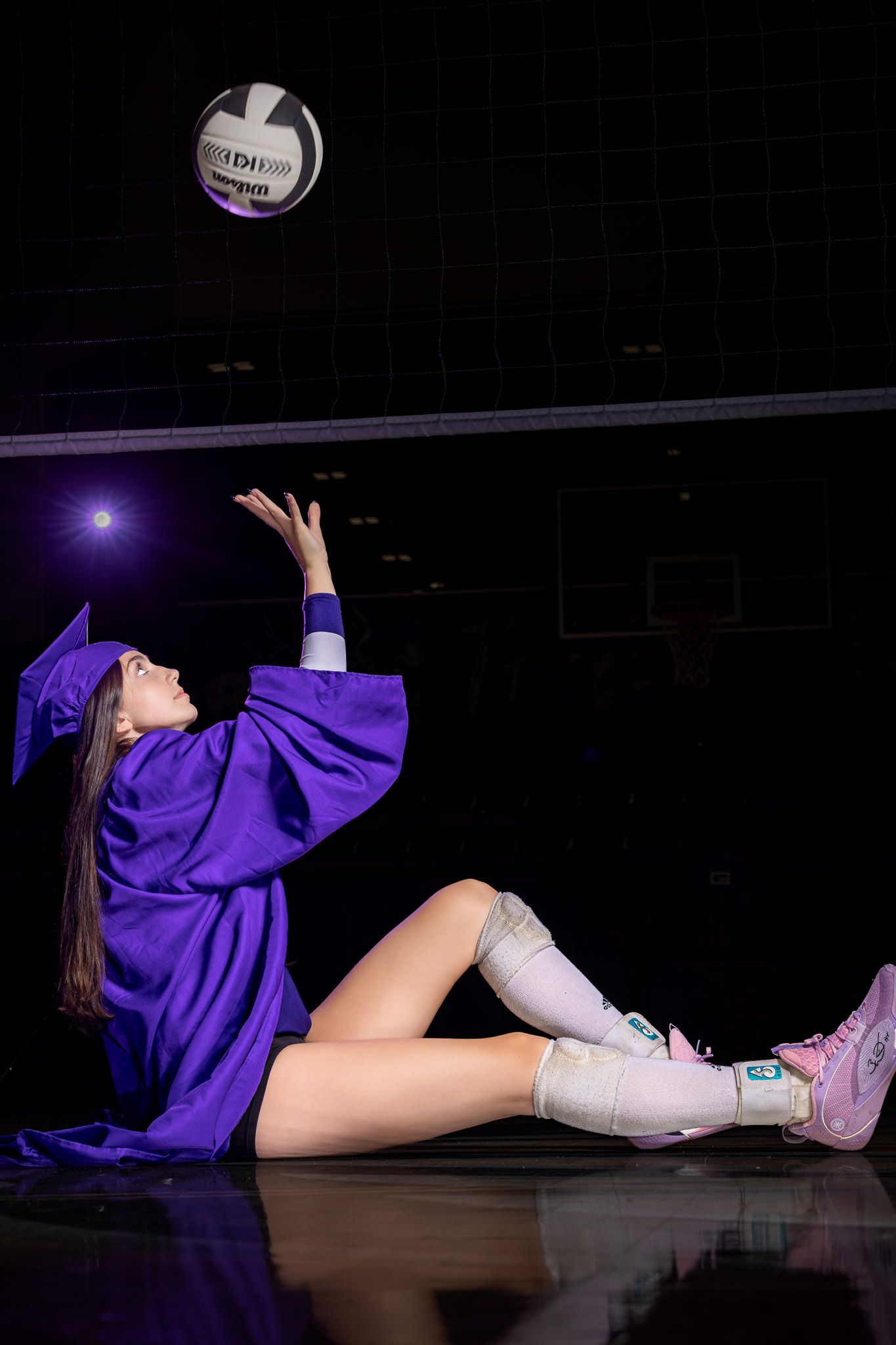A young woman in a purple graduation gown and cap tosses a volleyball while sitting on a gym floor.