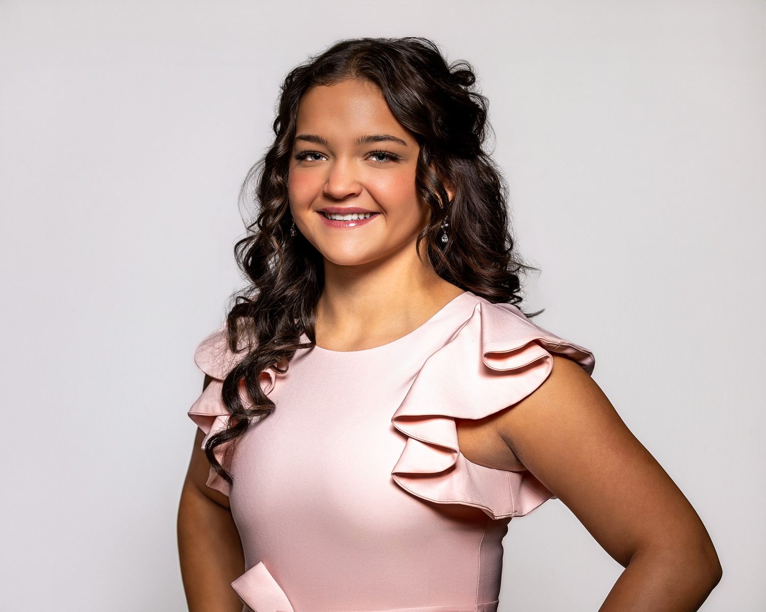 Young woman in a pink ruffled dress smiling confidently against a light gray background.