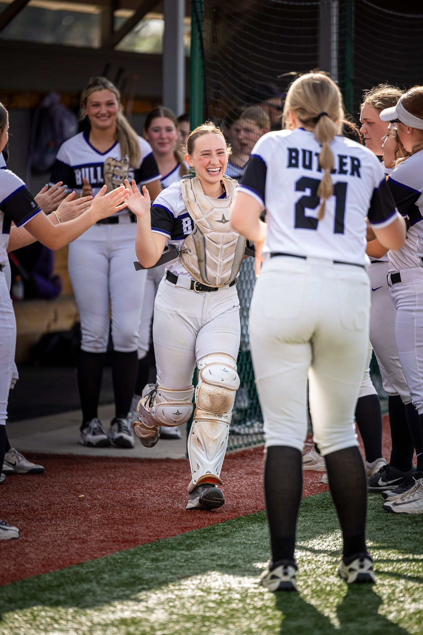 Smiling softball catcher in full gear walks past teammates giving high-fives while player Butler, number 21, stands in the foreground.