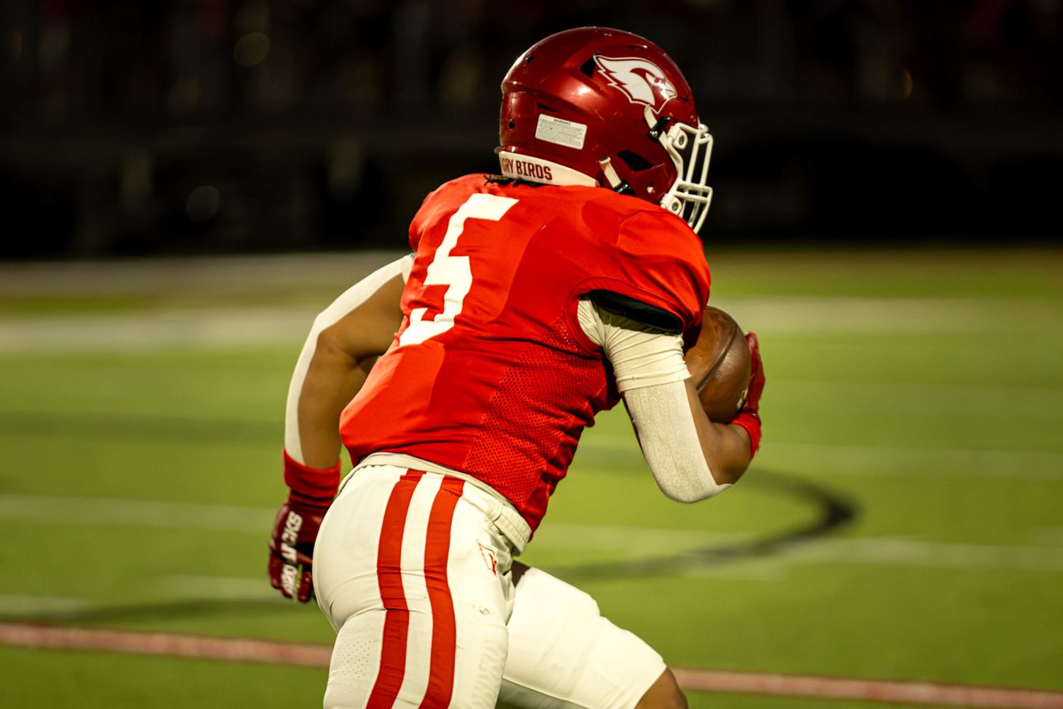 High school football player in a red jersey with number 5 and red helmet runs down the field carrying the football during a night game.