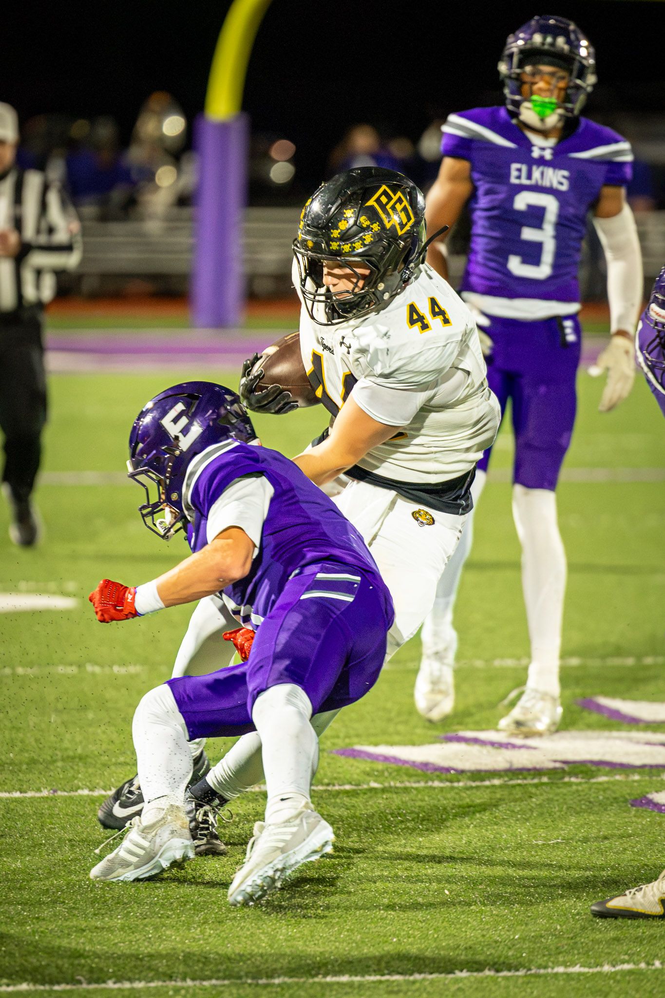 High school football player in white and gold jersey number 44 carries the ball while a purple-uniformed Elkins defender makes a tackle.