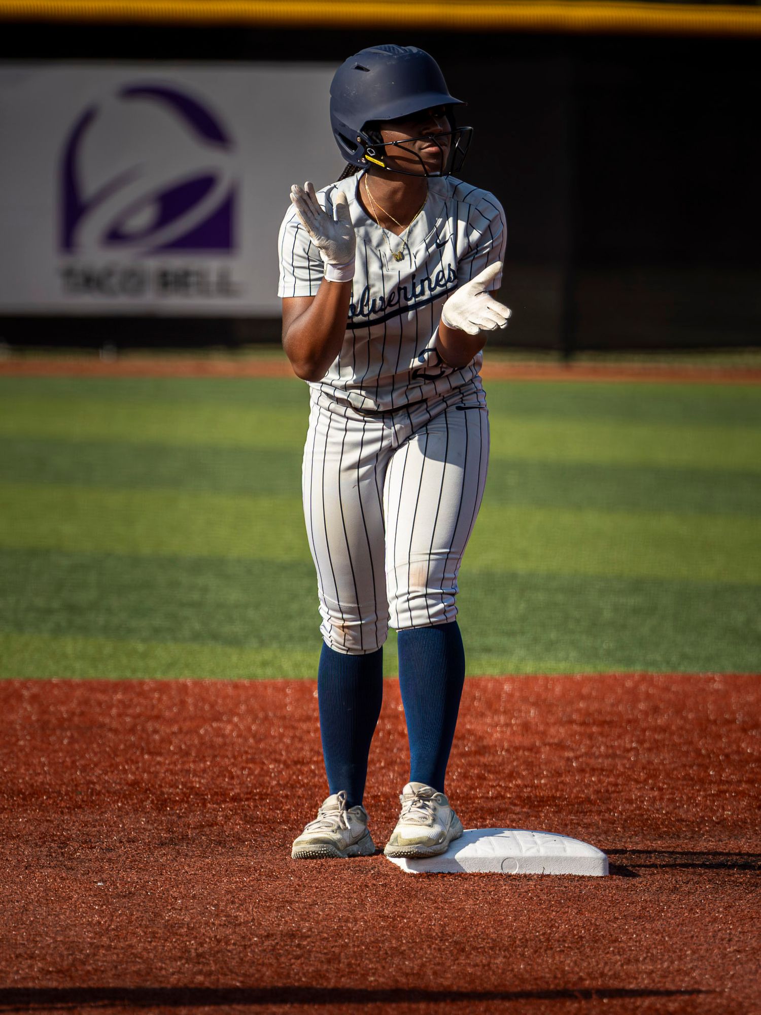 Softball player in a navy helmet and pinstripe uniform stands on second base, clapping her gloved hands on a sunny field.