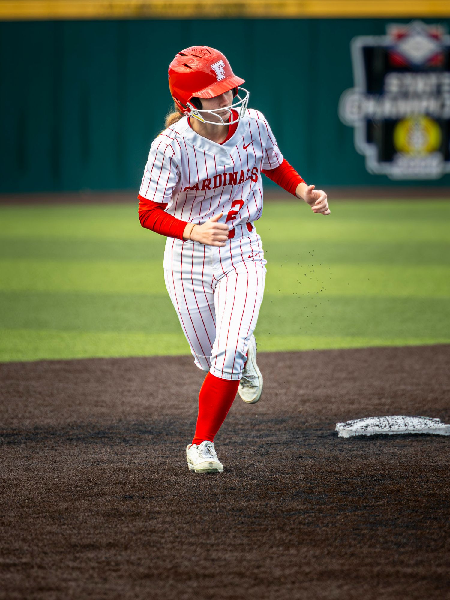 A softball player in a red helmet and white Cardinals pinstripe uniform runs the base paths on a dirt infield during a game.