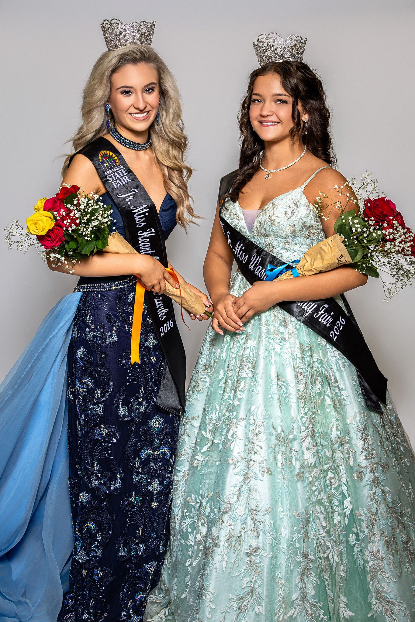 Two beauty queens in formal gowns wearing crowns and sashes hold flowers while posing together at a pageant.
