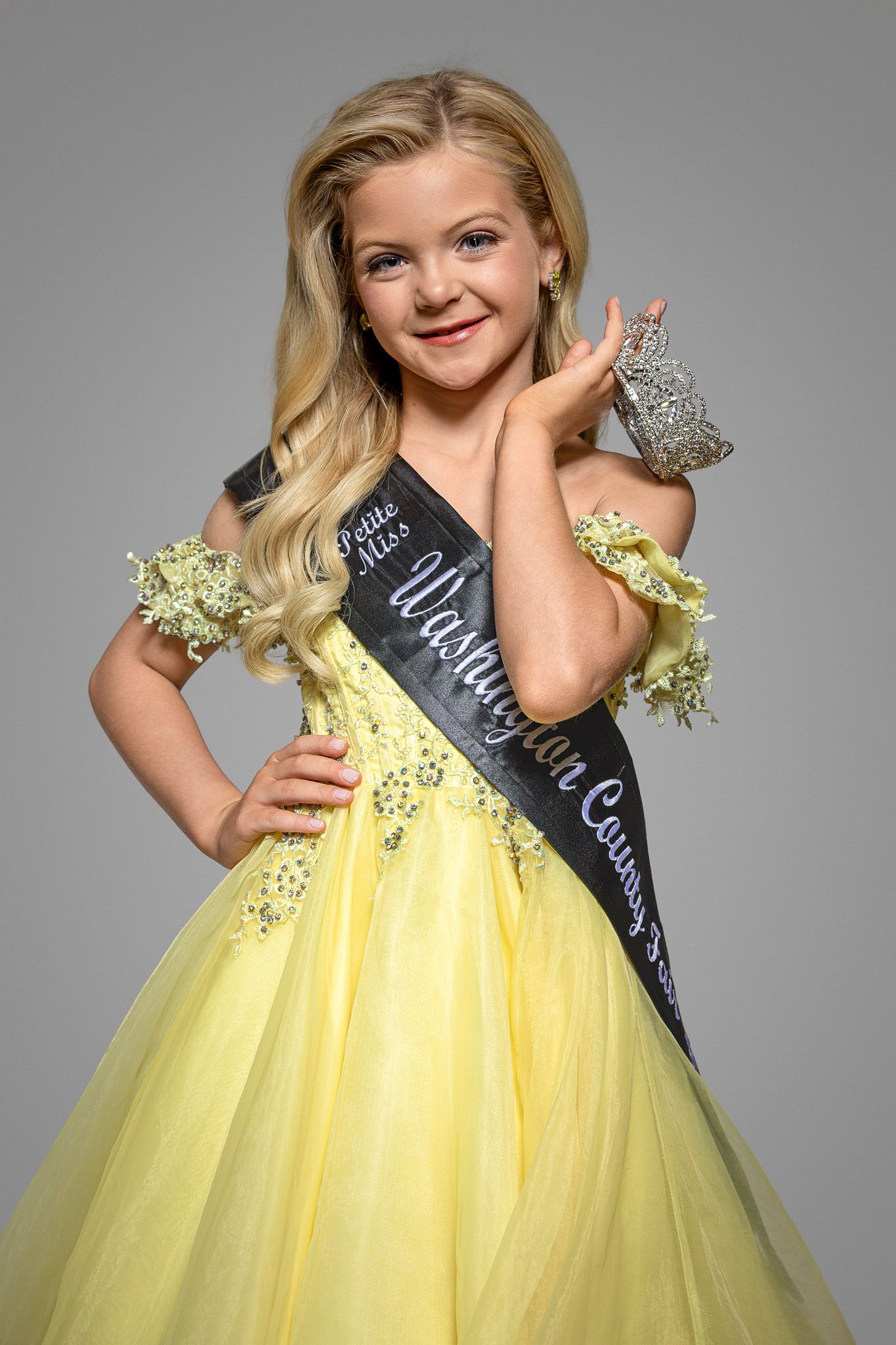 Young girl in yellow pageant gown wearing Little Miss Washington County sash and crown at a pageant.