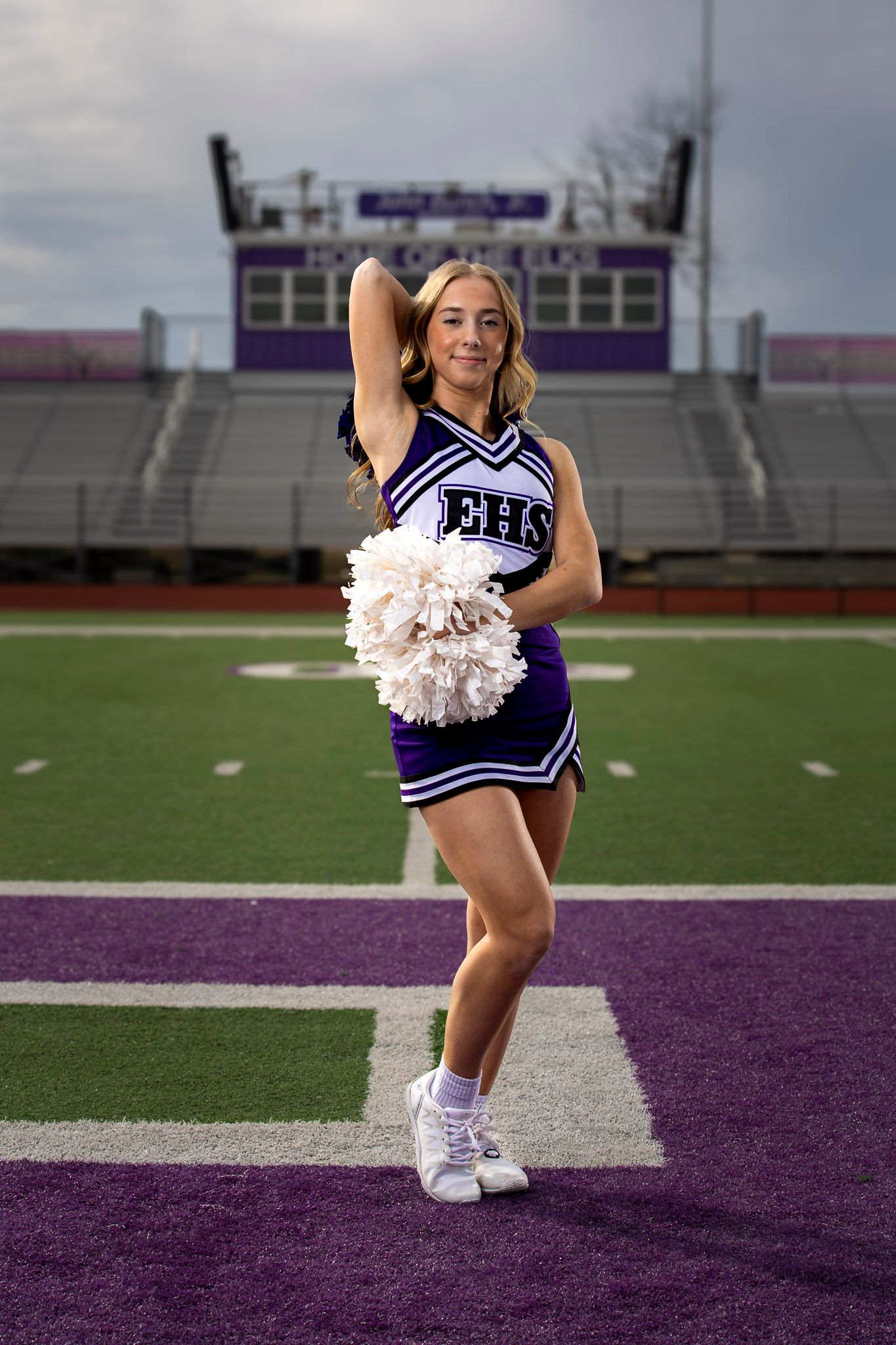 Elkins high school cheerleader in purple EHS uniform posing with pom-poms on football field stadium.