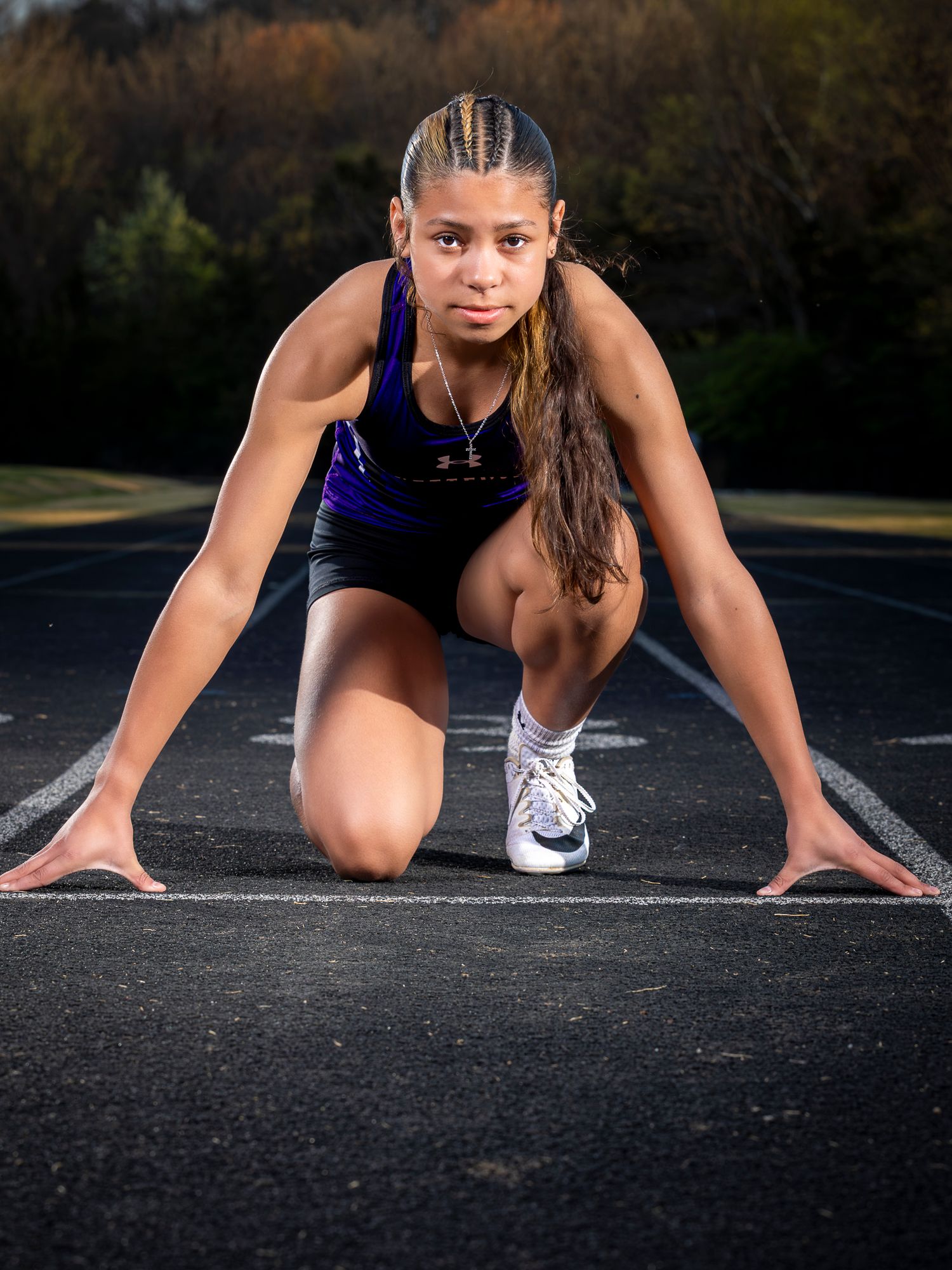 Young female sprinter in purple Under Armour uniform crouches in starting position on outdoor track, ready to race.
