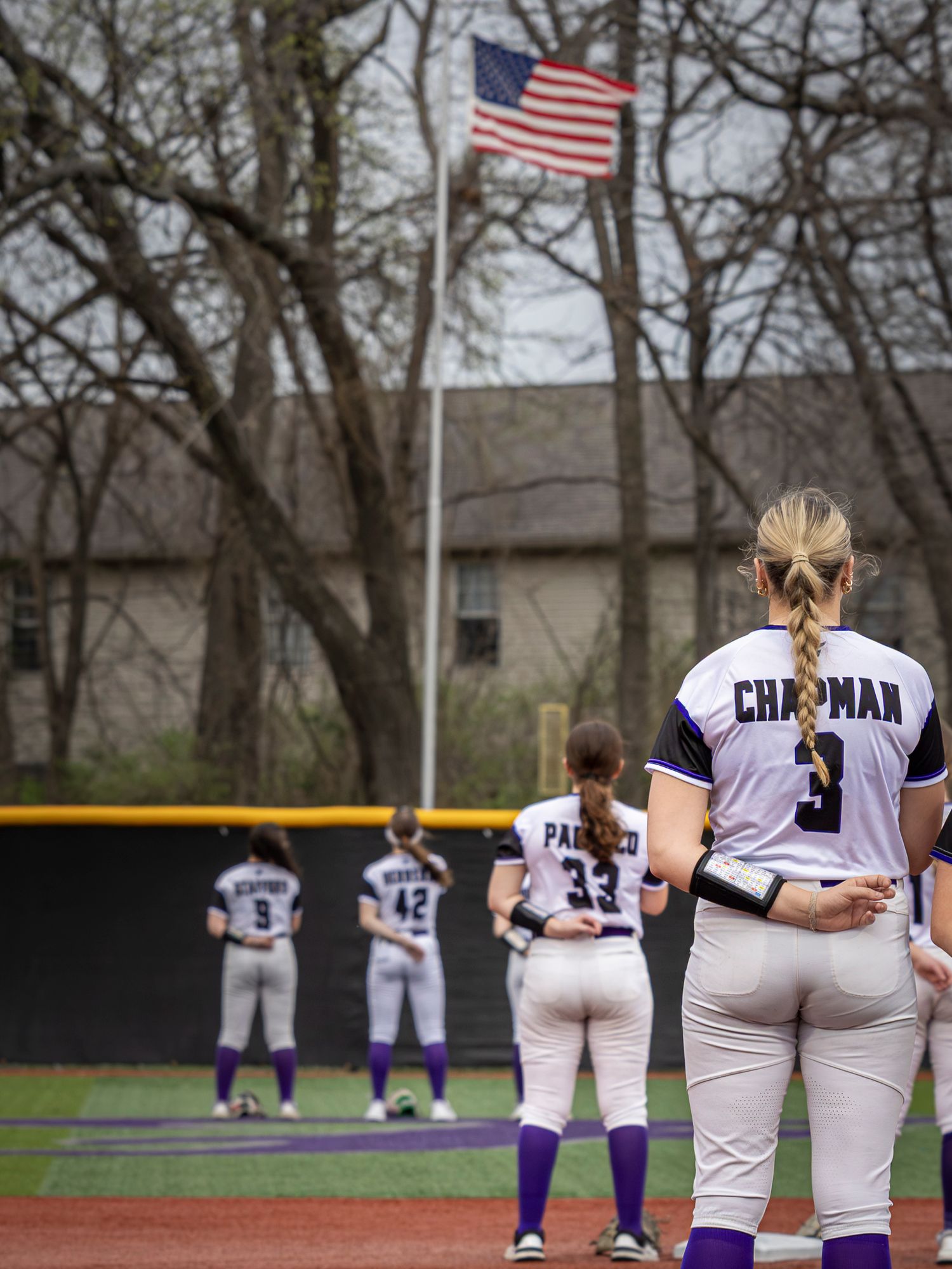 Softball players in white and purple uniforms stand at attention on the field facing an American flag on a pole beside bare trees.