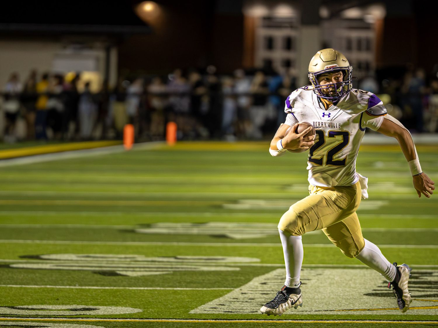 High school football player wearing number 22 sprints down the field at night while carrying the ball as spectators watch from the sideline.