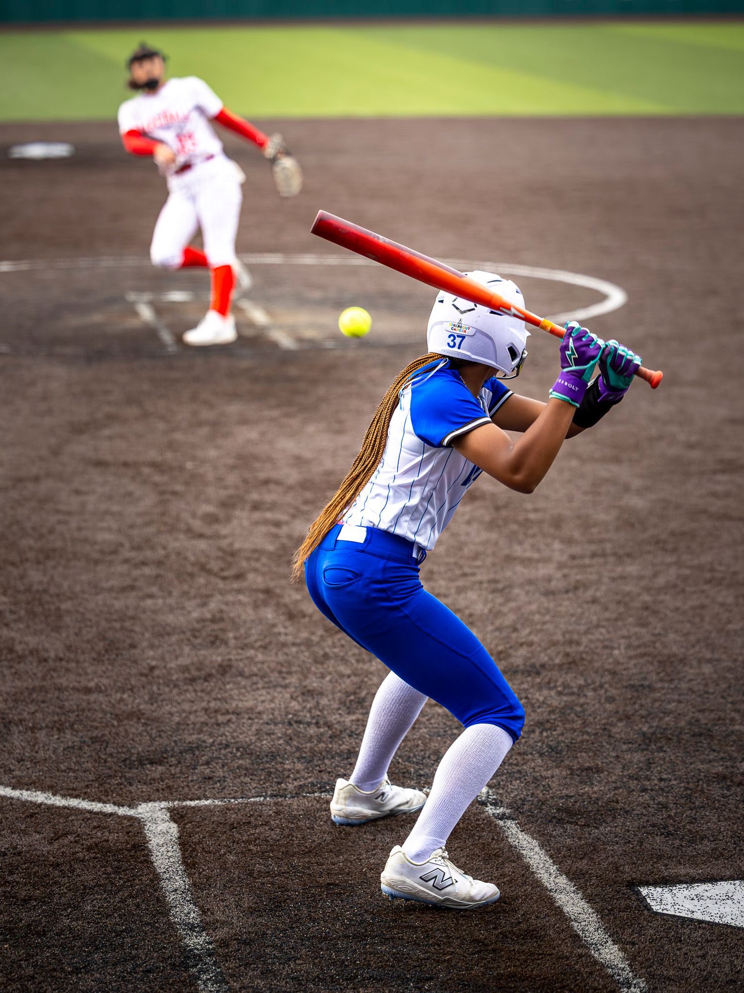 A softball batter in a blue and white uniform and helmet swings a red bat as the pitcher releases a yellow ball on the dirt field.