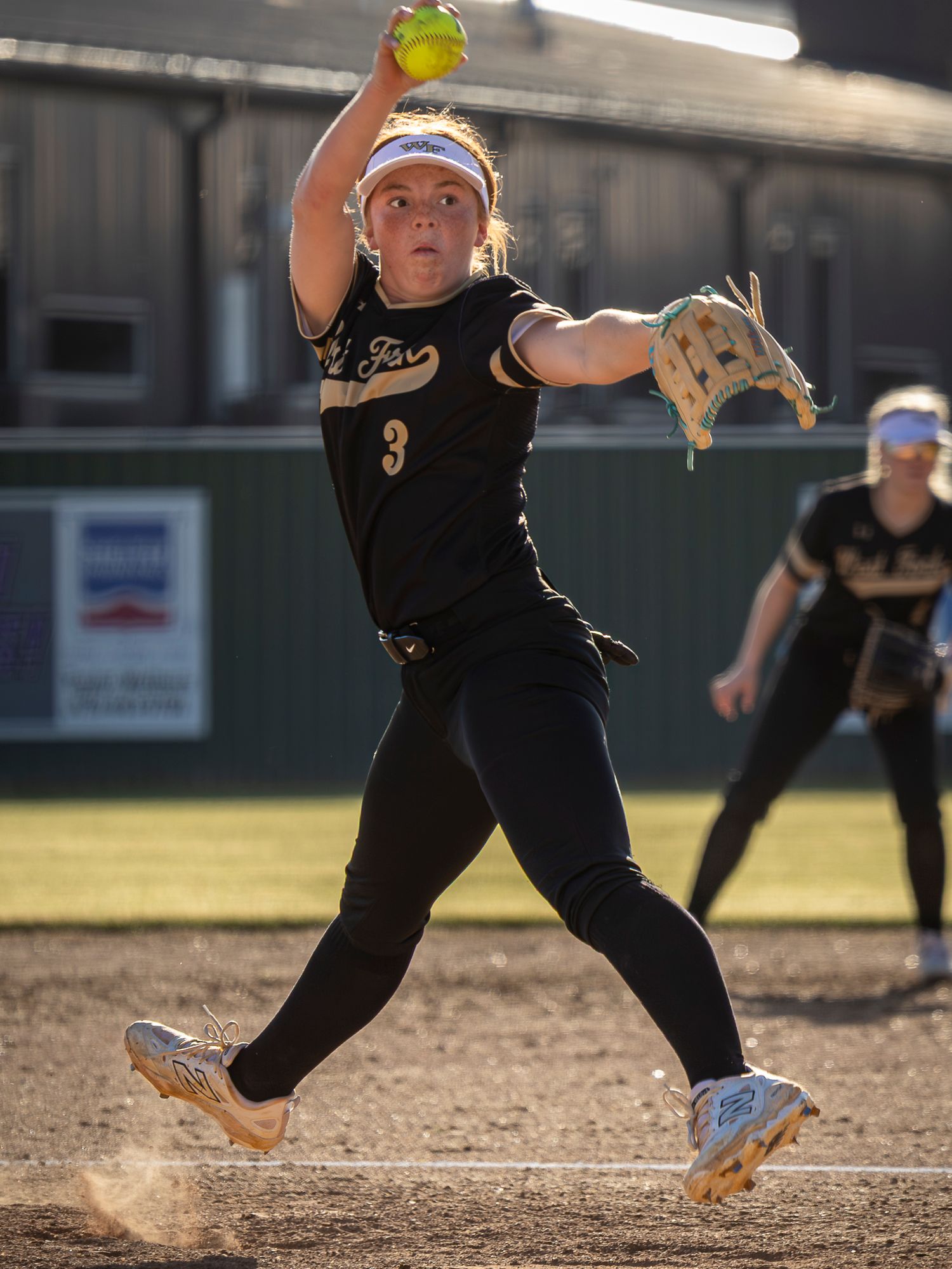 Softball pitcher in a black number 3 uniform and white visor winds up mid-pitch, holding a yellow softball during a game on the dirt.