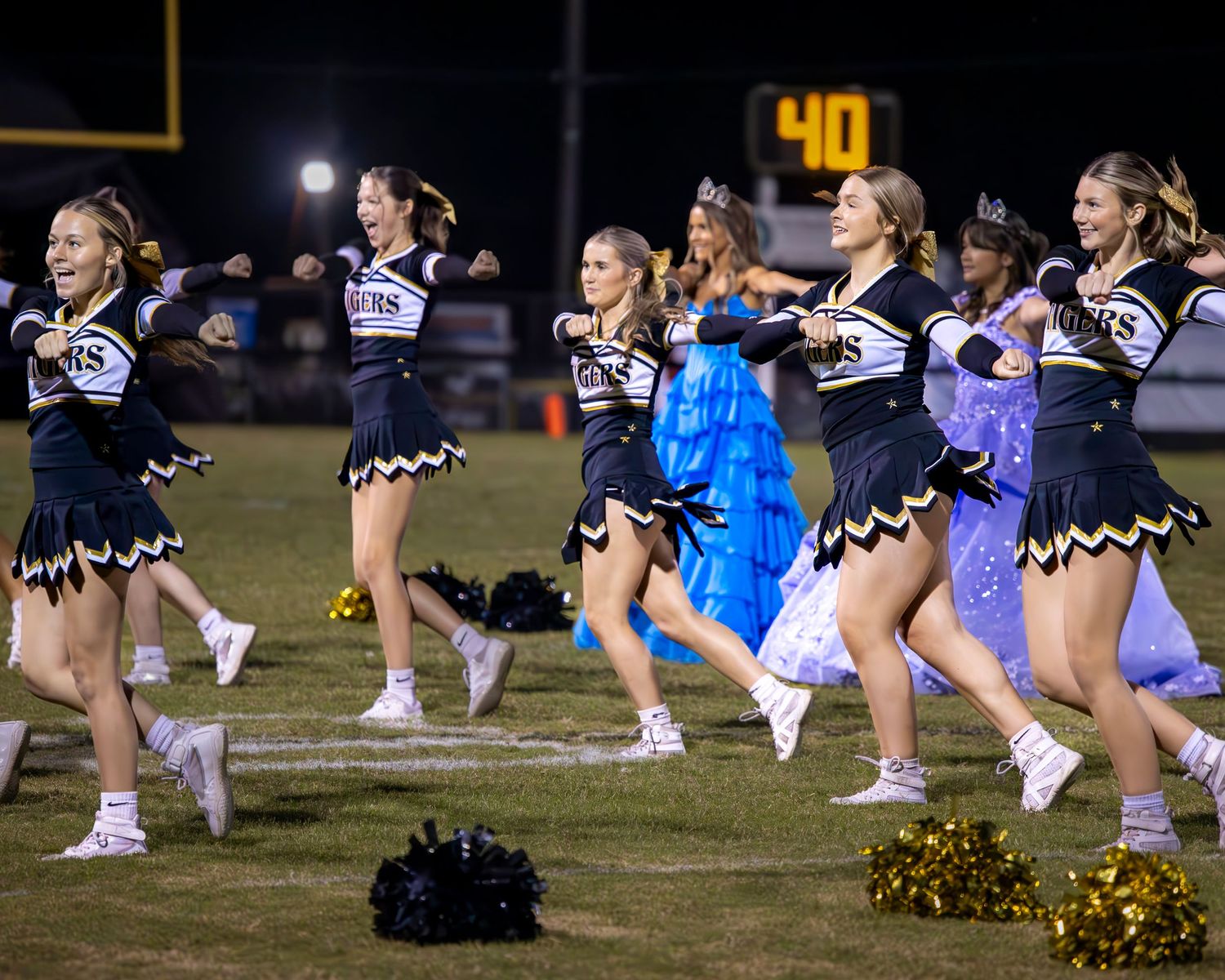 High school cheerleaders in black and gold uniforms perform energetic routine on football field at night game.