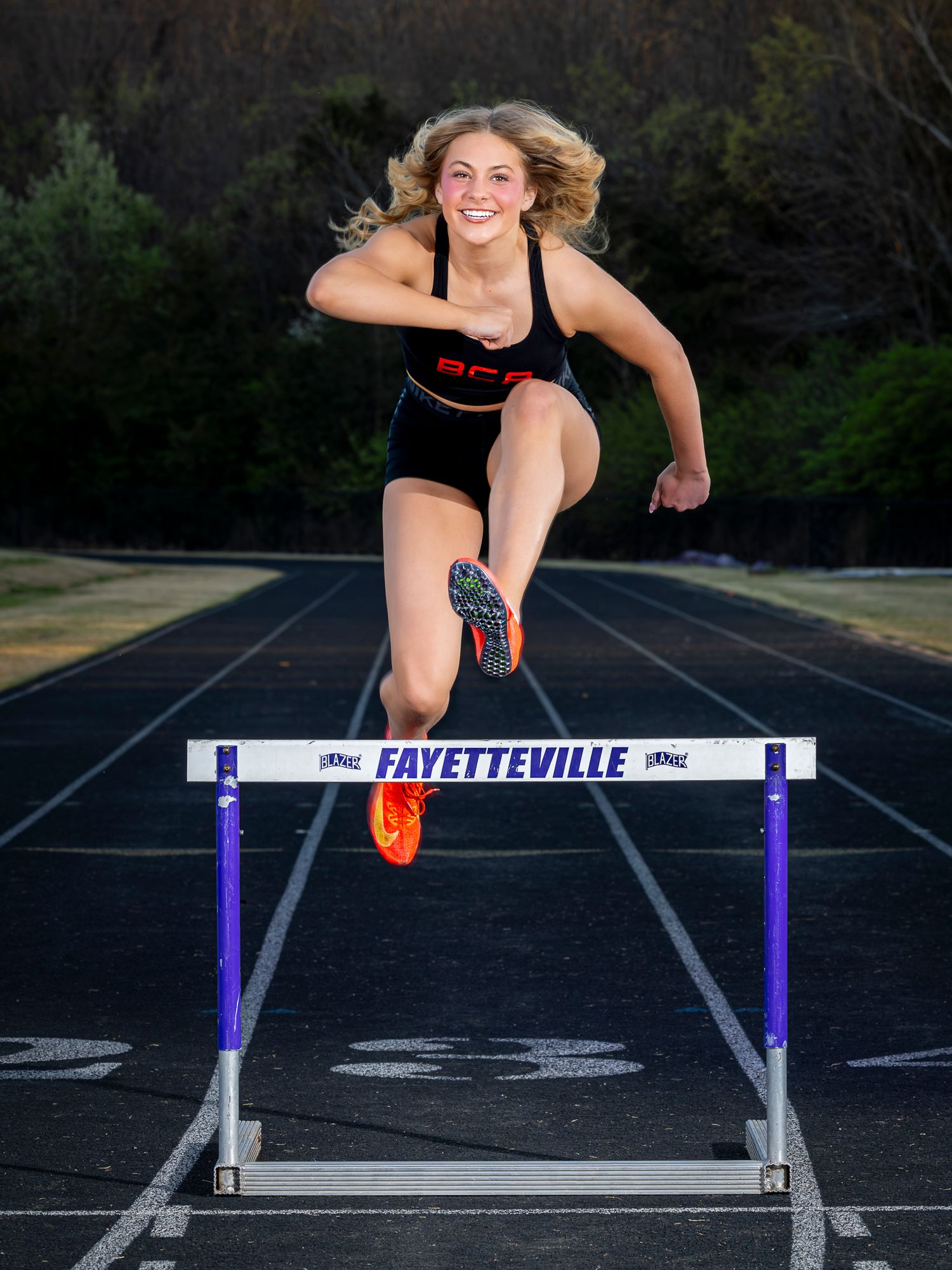 Female high school athlete leaping over a Fayetteville track hurdle in a black uniform, smiling mid-jump on an outdoor track.