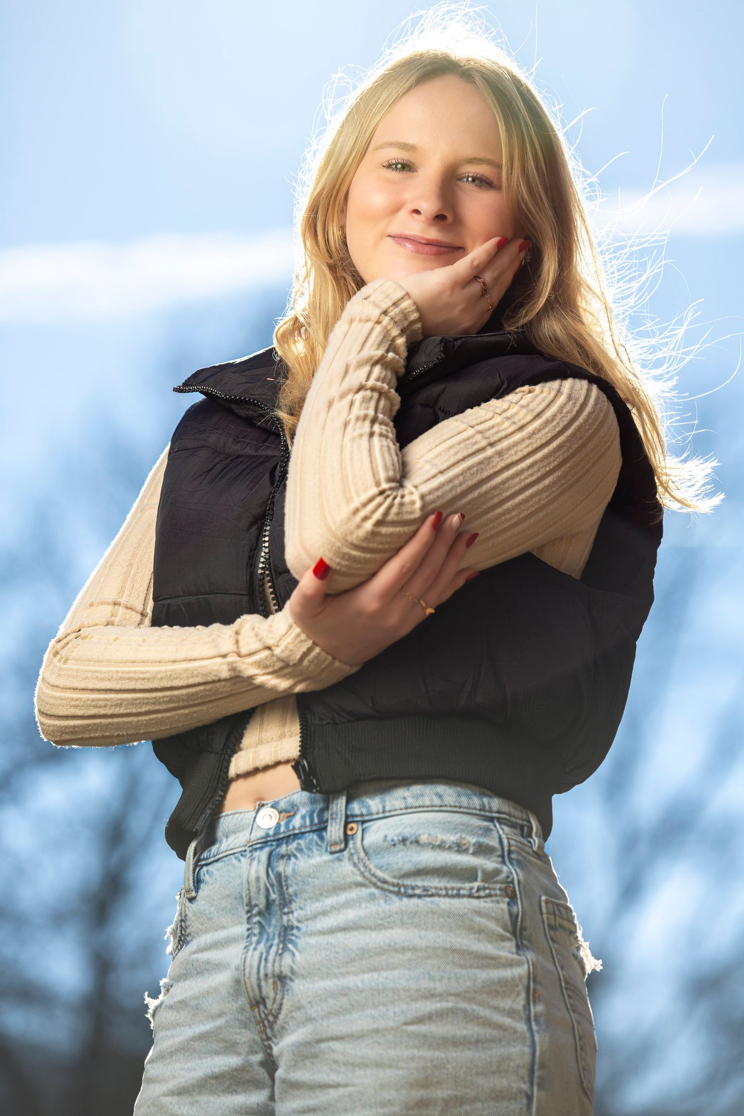 Young blonde girl in black vest and jeans smiles outdoors, hand on cheek, with blue sky background.