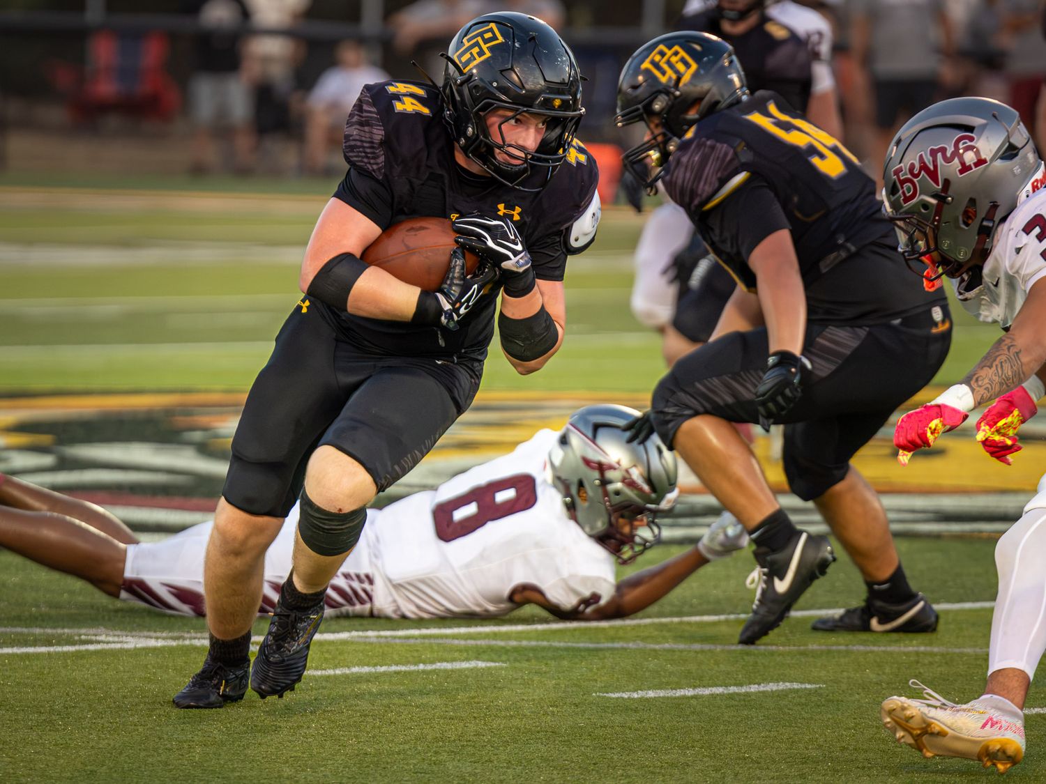 A high school football running back in a black and gold uniform carries the ball past a diving defender in a white jersey on the field.