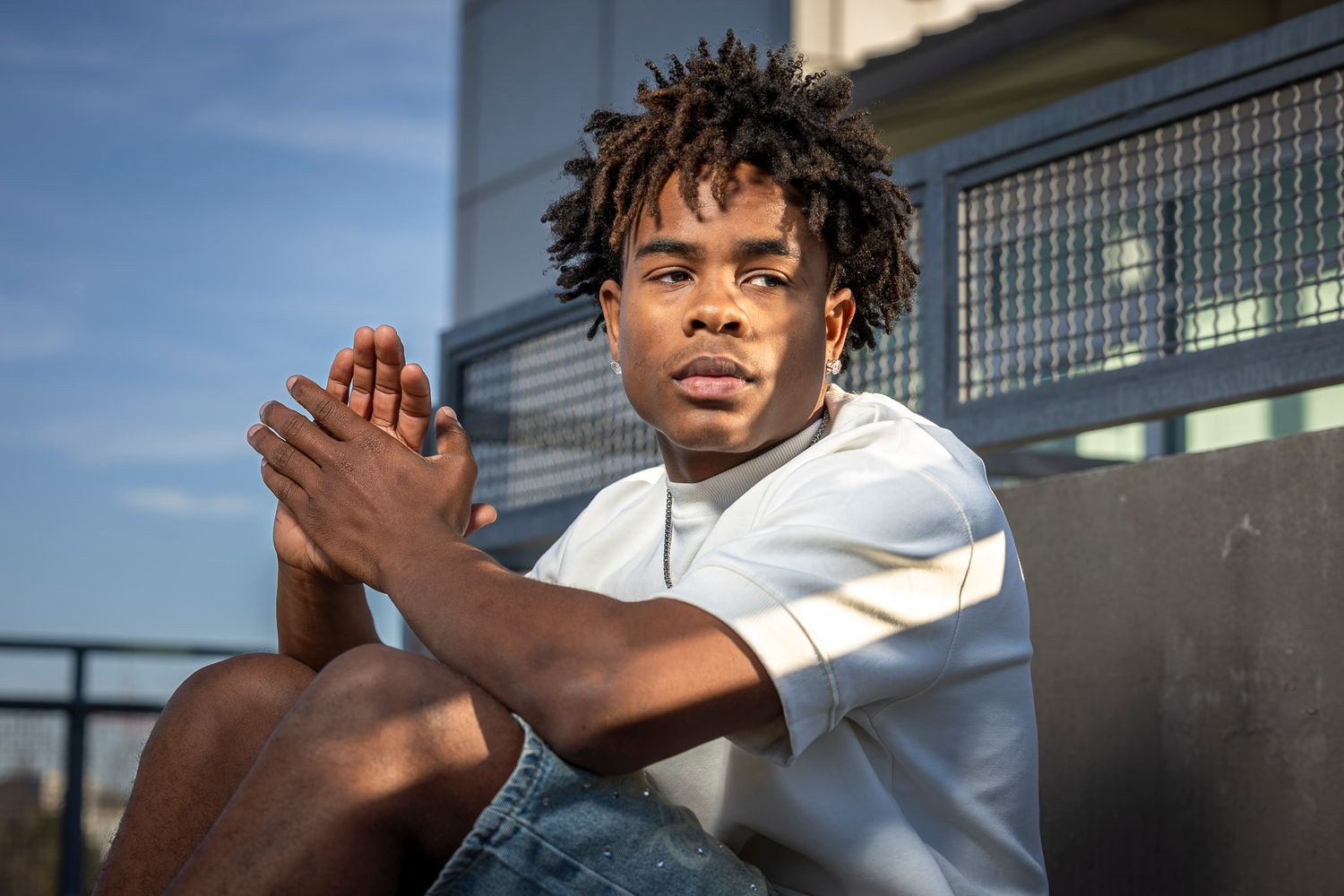 Young Black man with locs sits outdoors in white tee and jeans, hands clasped, looking confidently at camera.