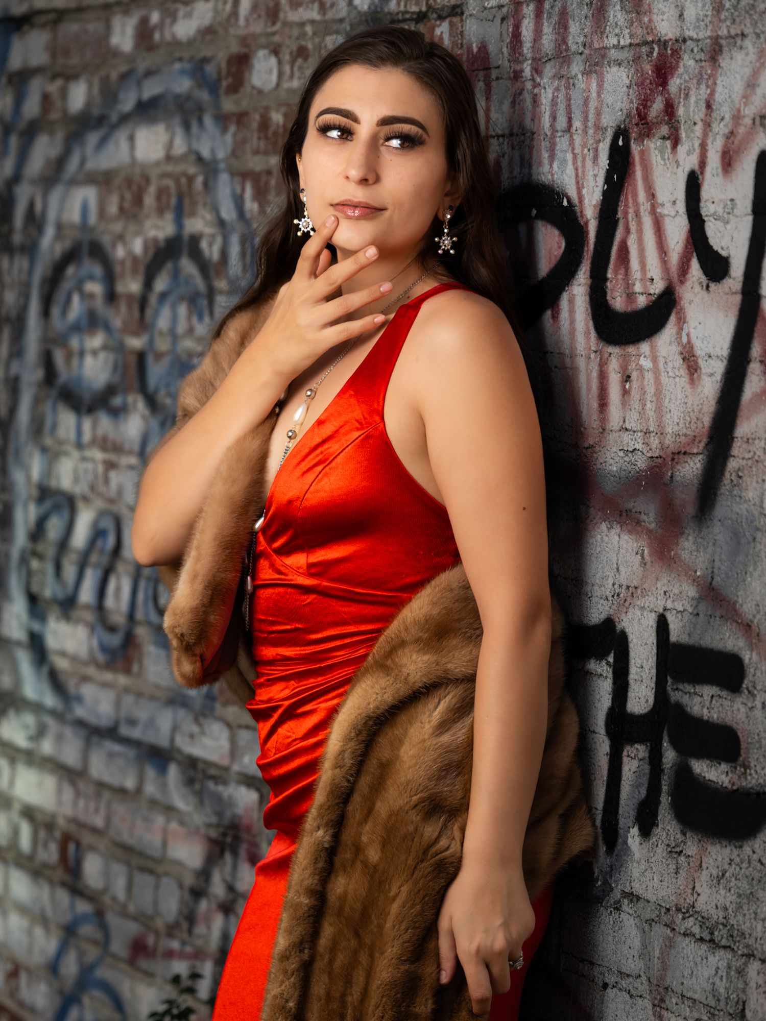 Woman in red satin dress with fur stole poses against graffiti wall, wearing chandelier earrings.