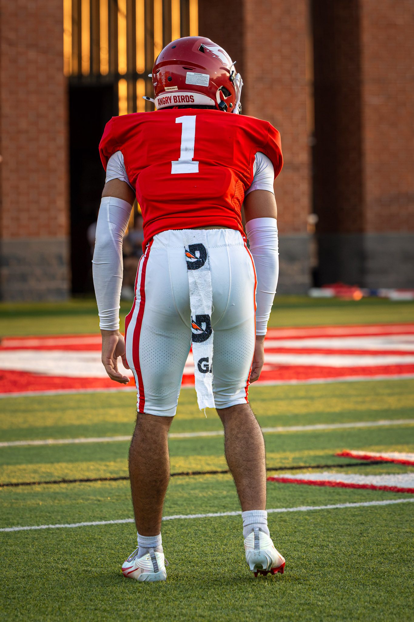 Football player in a red number 1 jersey and helmet stands in a ready stance on a turf field with a play call sheet on his pants.
