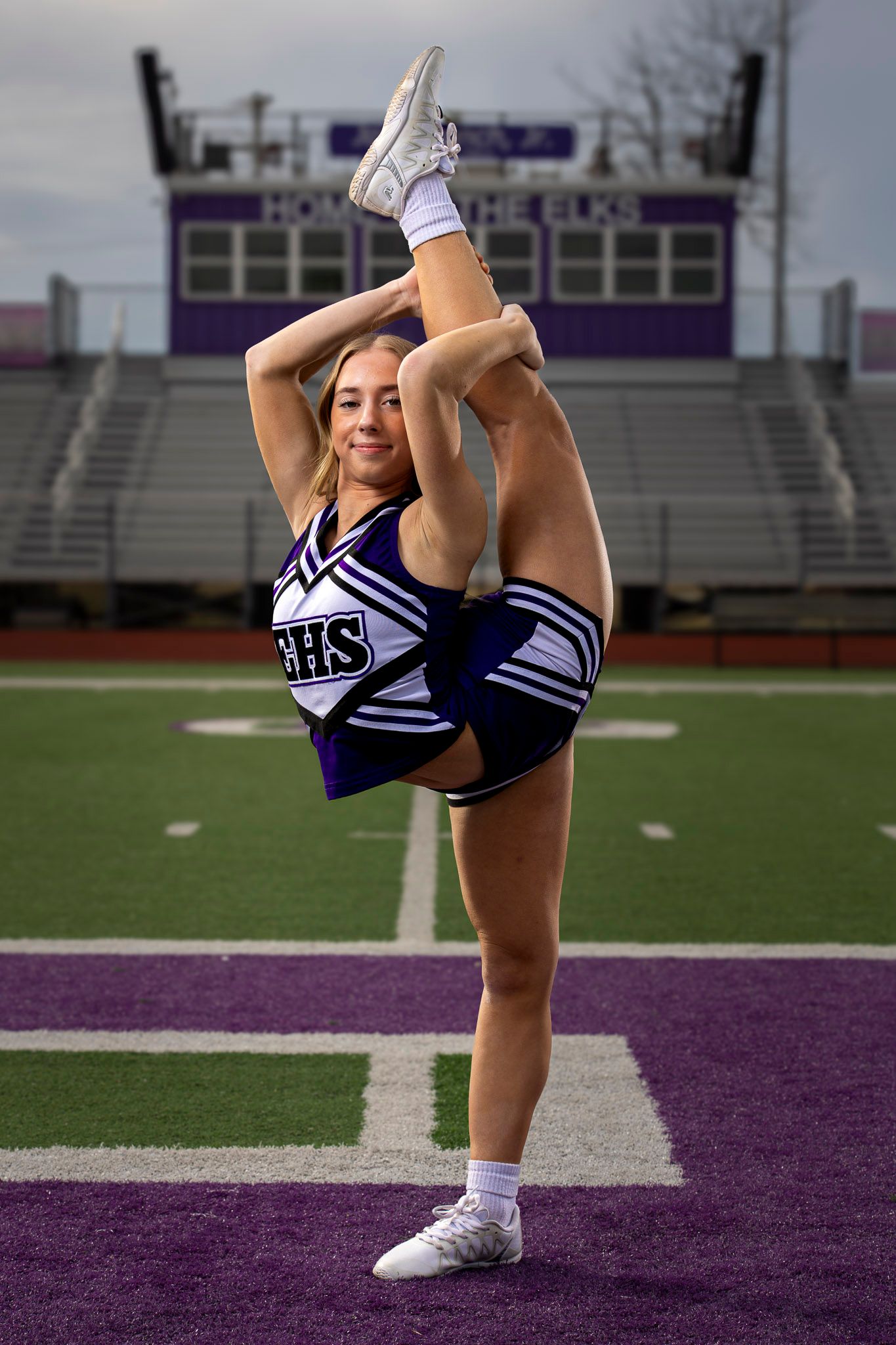 Cheerleader in a purple and white uniform performs a standing scorpion stretch on a purple and green football field with bleachers behind.