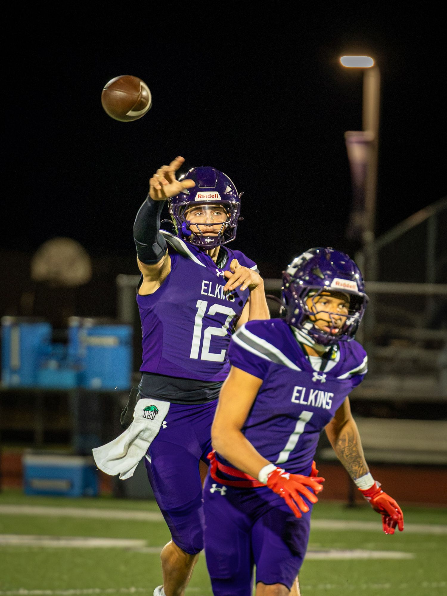 Elkins football quarterback in purple jersey number 12 throws a pass as teammate number 1 runs past during a night game.