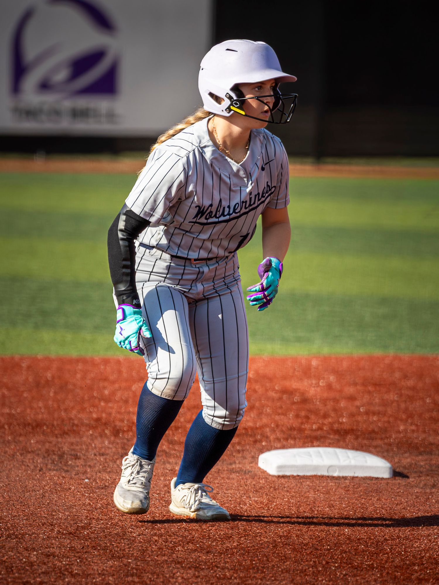 Softball player in a pinstriped Wolverines uniform, helmet and batting gloves leads off second base on the red dirt infield.