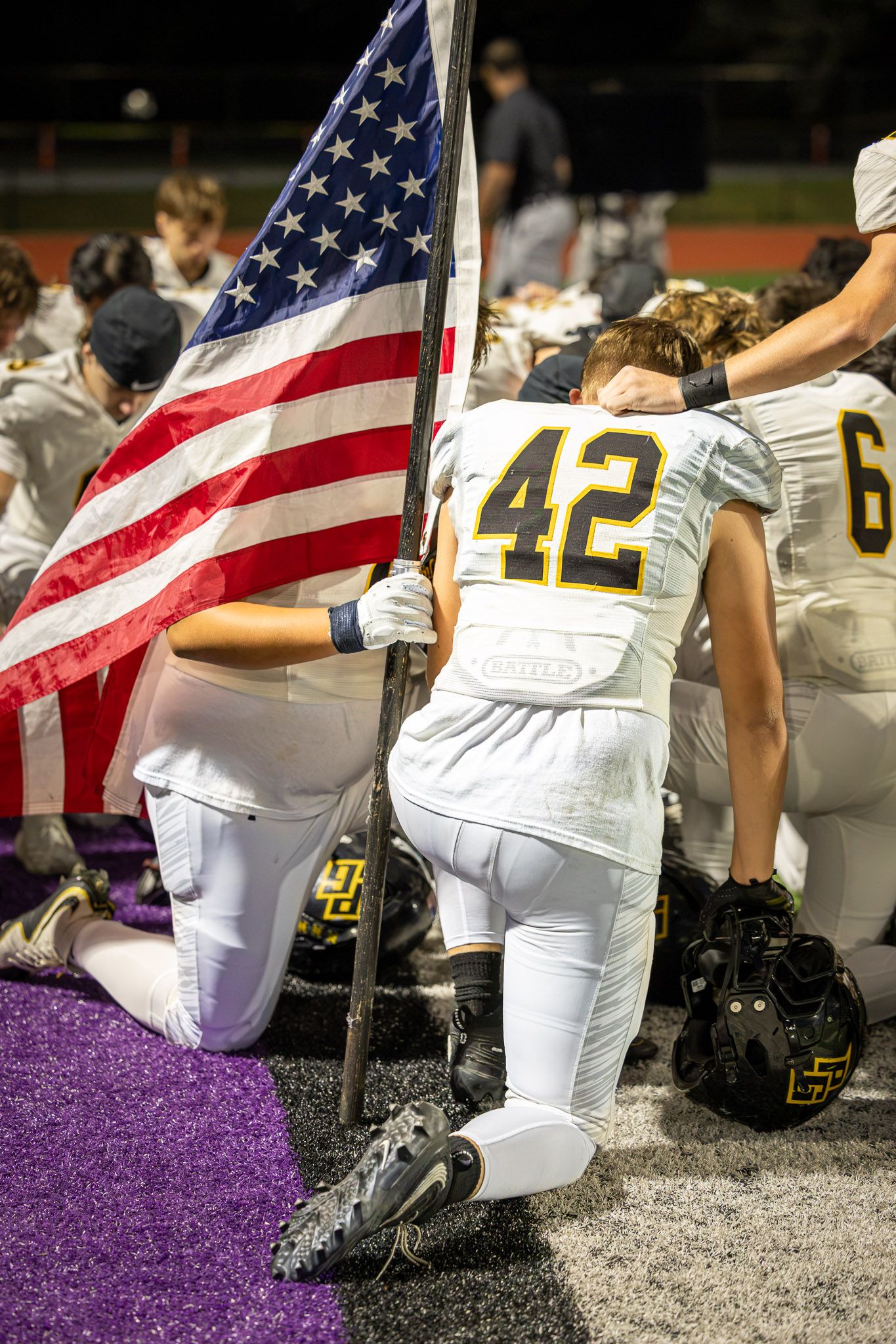 Football player in white jersey number 42 kneels on the field holding an American flag while teammates kneel in prayer beside him.