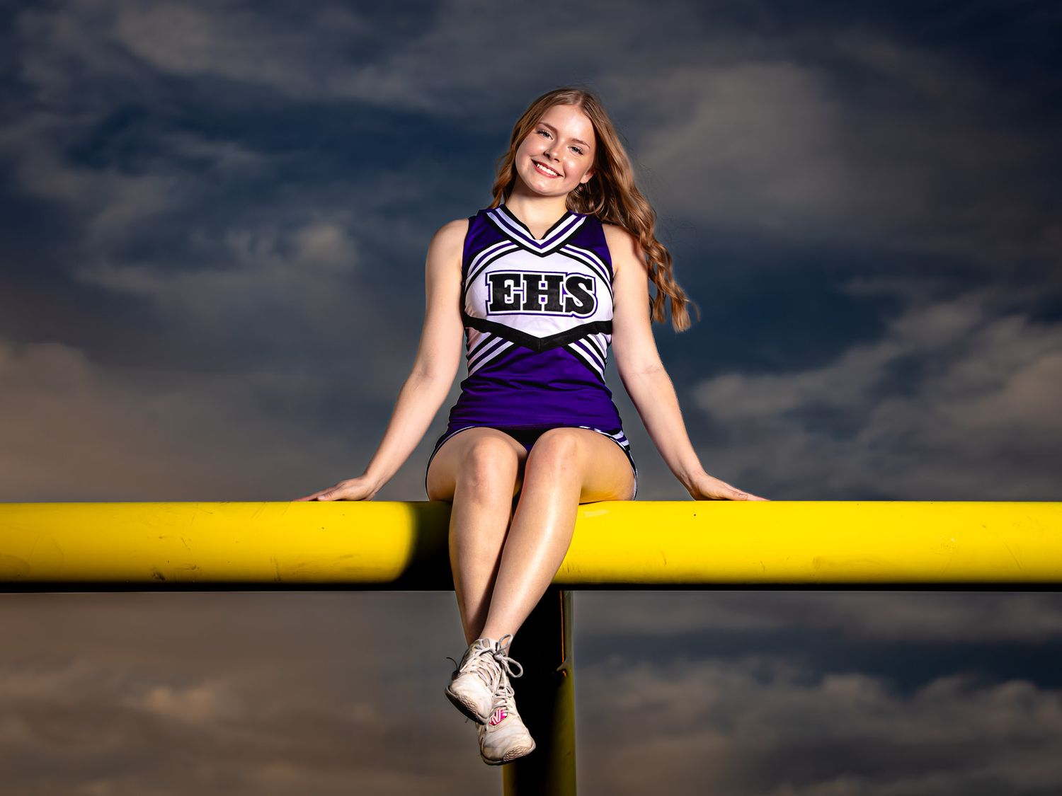 Cheerleader in purple EHS uniform sits on yellow railing against dramatic cloudy sky.