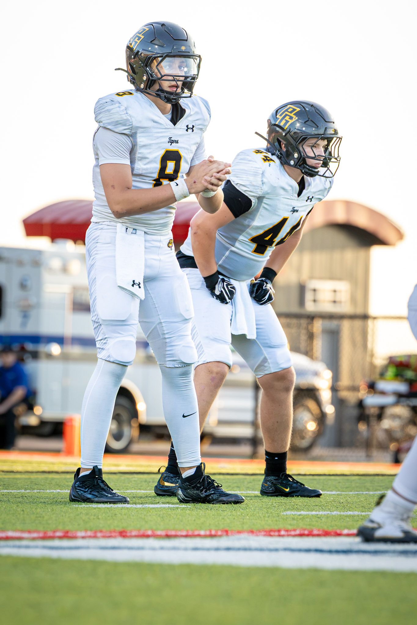 Two football players in white jerseys and black helmets stand ready in a pre-snap stance on a grass field during a high school game.