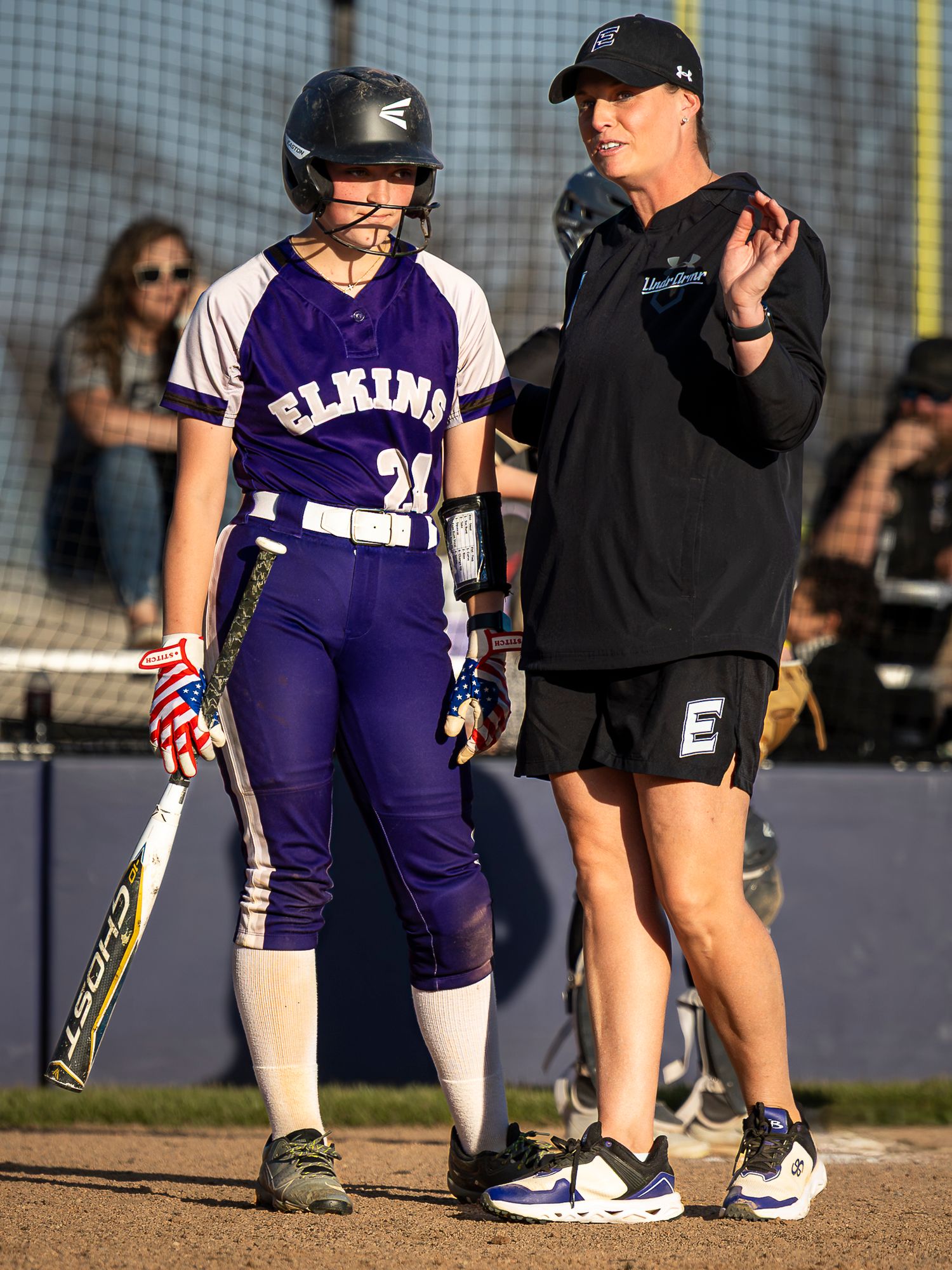 Elkins softball player in a purple uniform and batting helmet holds a bat while a coach in black gestures beside her on the field.