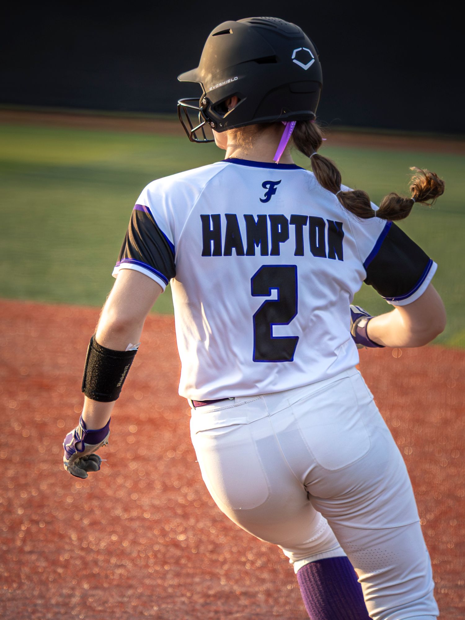 Softball player in a white Hampton number 2 jersey and black helmet with braided hair runs along the base path on a dirt infield.